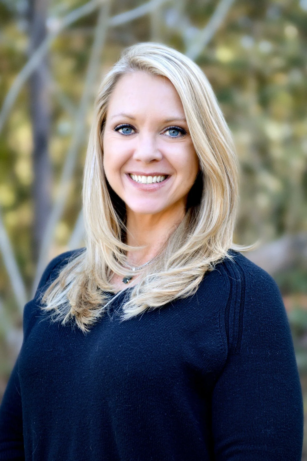 Portrait of a smiling woman with blonde hair and blue eyes, wearing a black top, outdoors with blurred trees in the background.