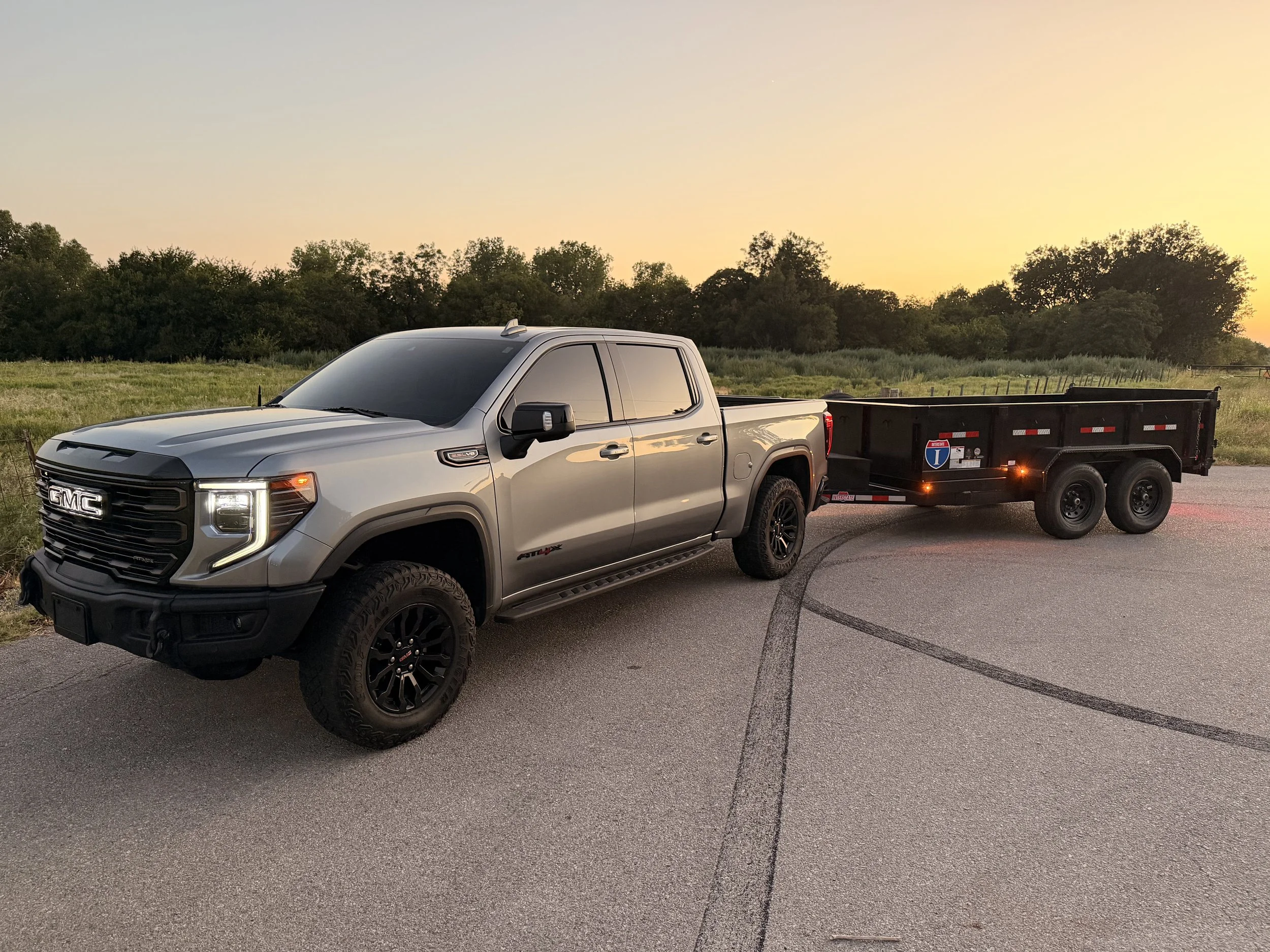 A silver GMC pickup truck towing a black utility trailer on a paved road during sunset, with green fields and trees in the background.
