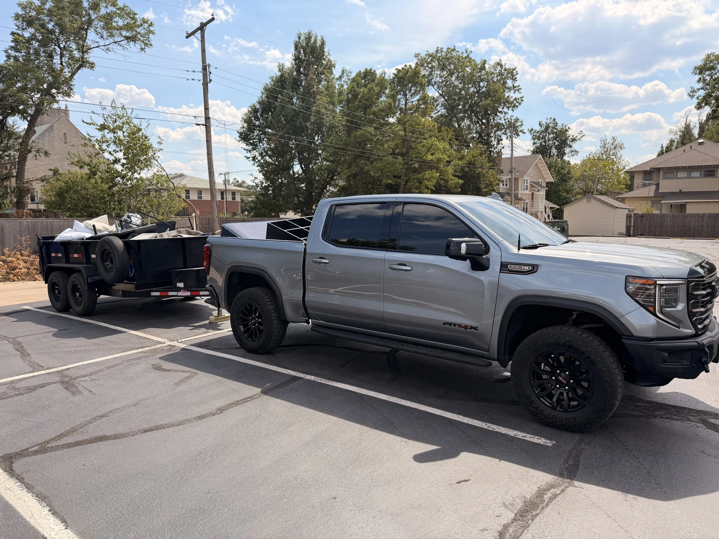 A gray pickup truck parked in a parking lot with a trailer loaded with various items hitched to it. The background shows trees, houses, and a partly cloudy sky.