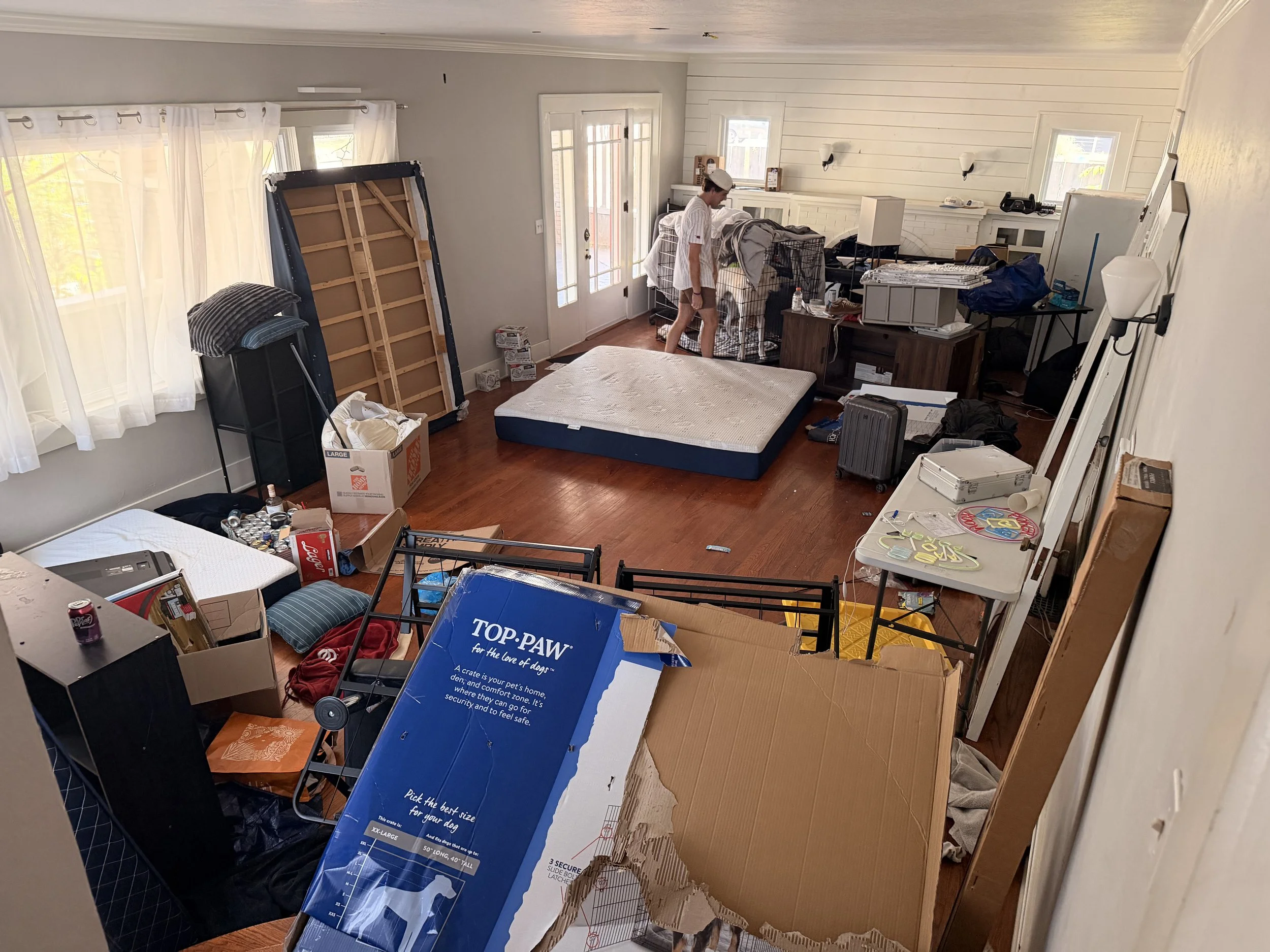 A cluttered living room with various furniture and items, including a mattress, boxes, and pet cage, with a person organizing in the background.