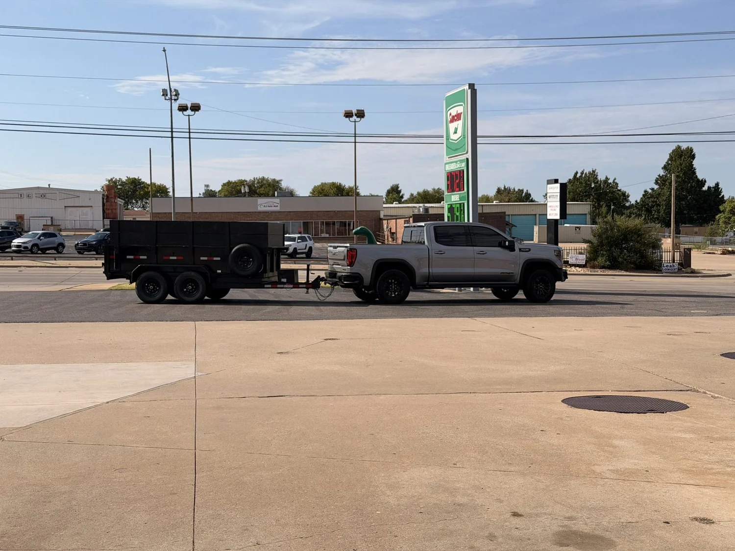 Side view of the Nicks Junk Removal silver pickup truck towing a black 6x12 dump trailer, parked at a gas station