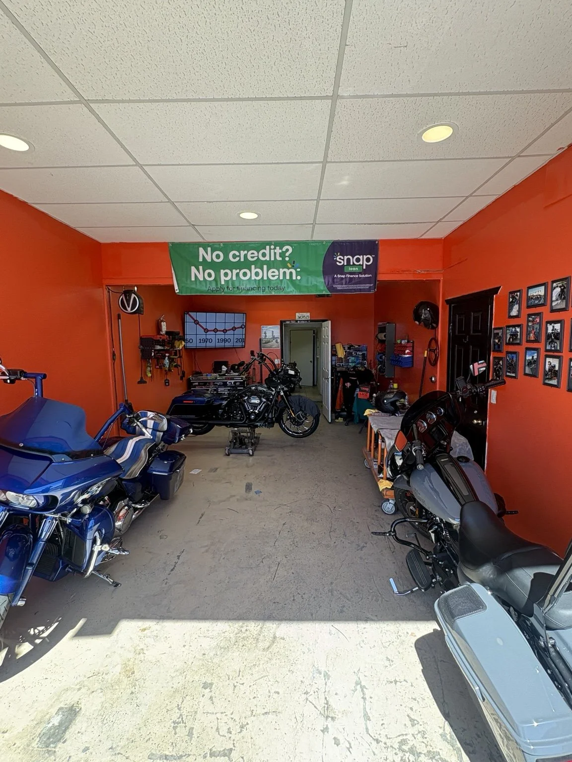 Interior of a motorcycle repair shop with multiple motorcycles parked inside and a green banner with white text that reads 'No credit? No problem'. The shop has red walls, framed pictures on the right wall, and various tools and equipment.