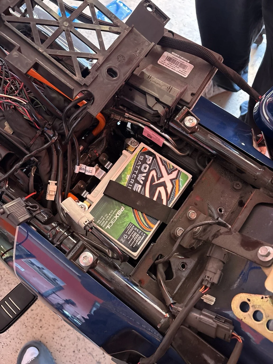 Close-up of an exposed car engine compartment showing a Power Zone battery secured with a strap, various wires, and mechanical parts.