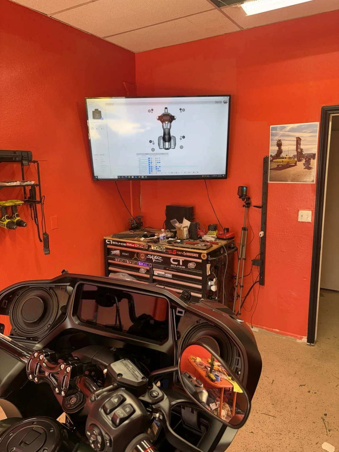 A workshop with a motorcycle in the foreground and a computer setup in the background. The computer screen displays a 3D model of a motorcycle. The workshop has orange walls, a tool drawer, and various tools and equipment on the side and on the desk.