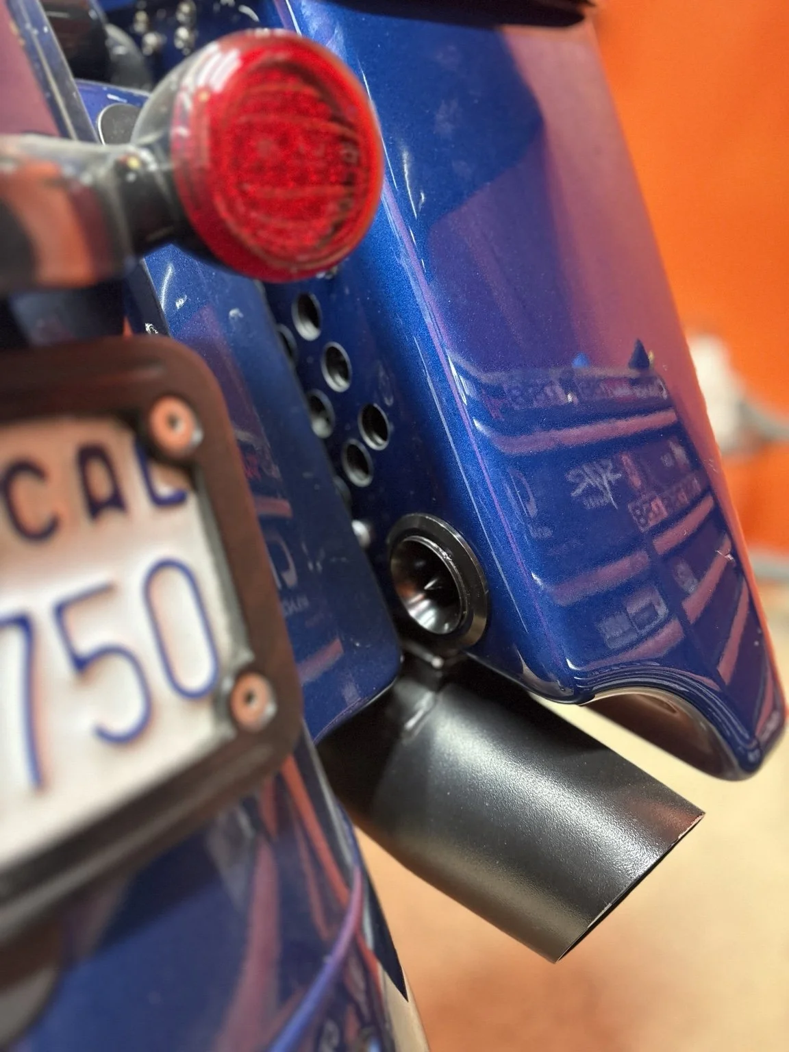 Close-up of a blue motorcycle's rear section showing a red tail light, a part of the license plate, and exhaust pipe.