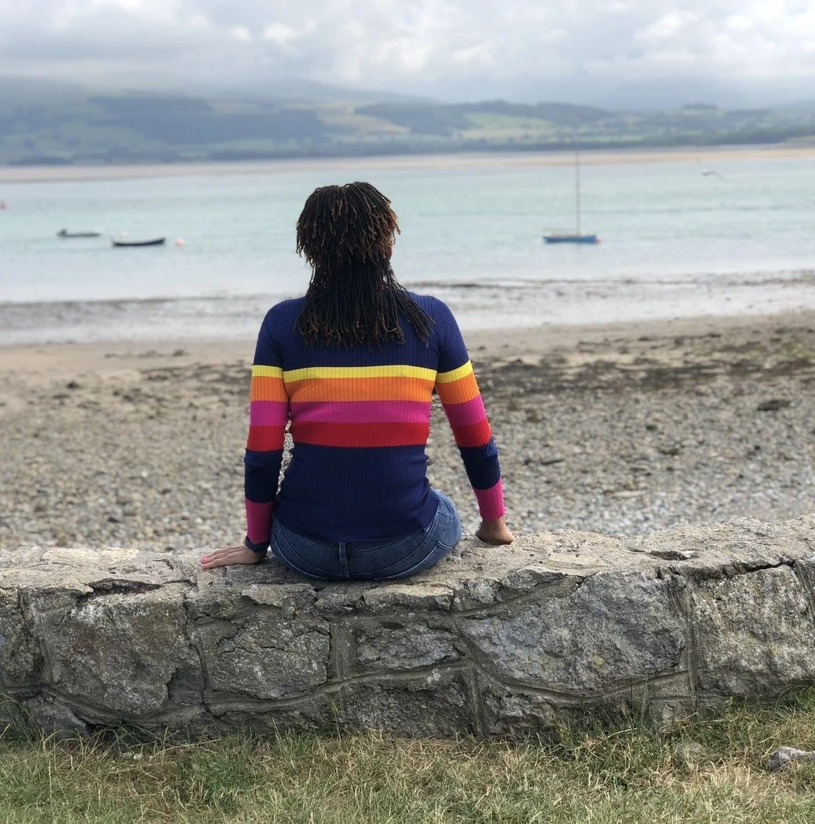 A woman with curly hair sitting on a stone wall at the beach, facing the water with sailboats in the distance, wearing a colorful striped sweater and jeans.