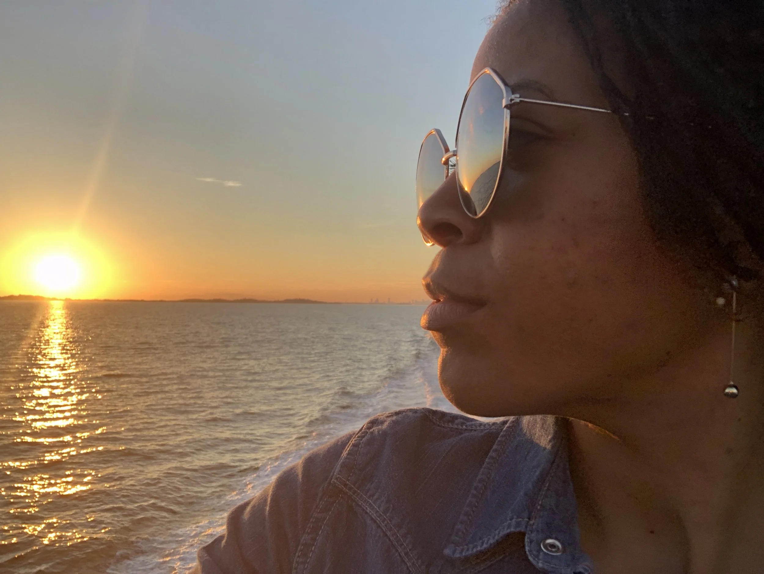 A young woman is wearing sunglasses and looking at a sunset over the water. Her face is in profile, and she appears to be near the shore, with the sun setting on the horizon.