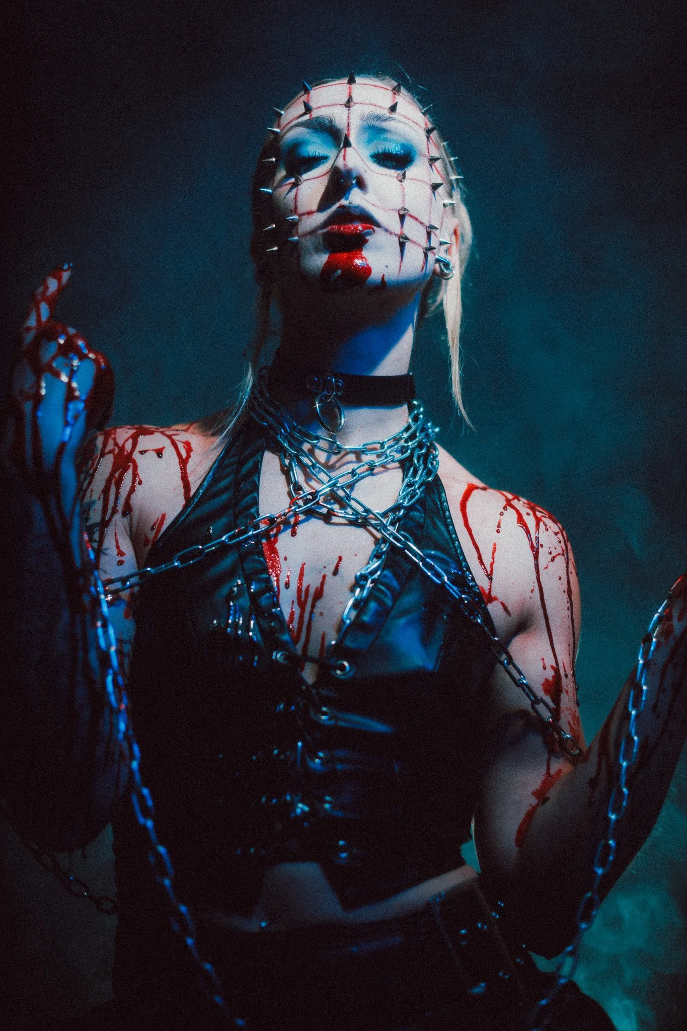 A woman with platinum blonde hair and dramatic makeup, wearing a black leather outfit, chains, and with blood splattered on her skin. She has a headpiece with spikes, red lipstick, and light blue eyeshadow, in a dark, moody setting.