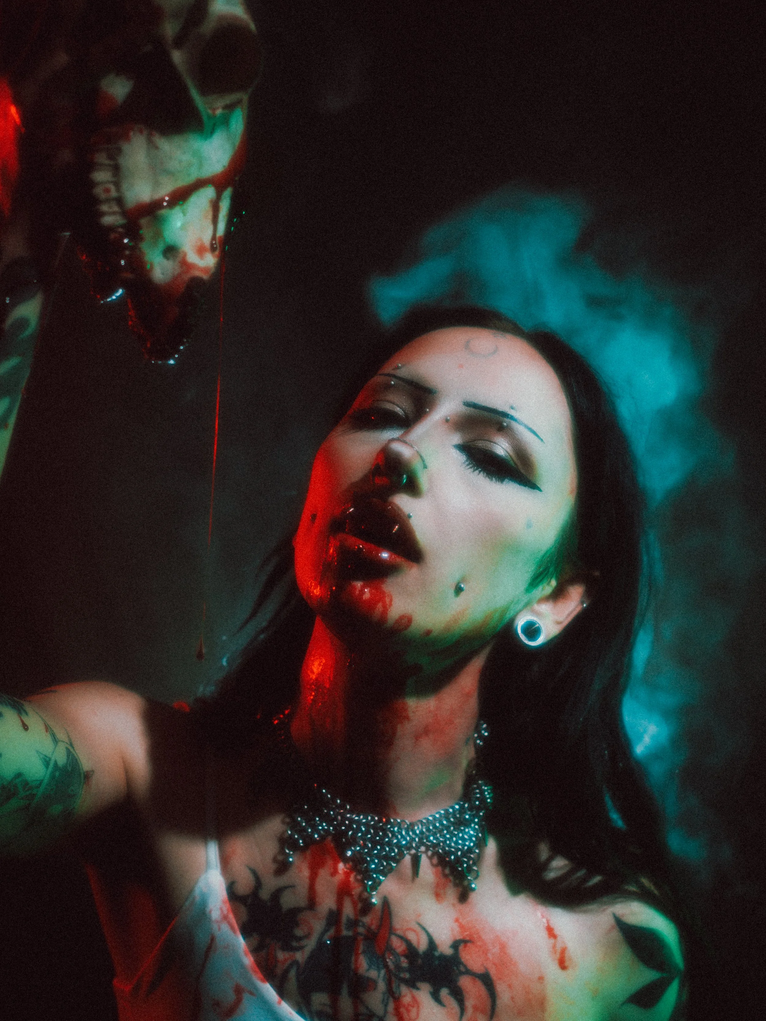 A woman with dark hair, tattoos, and piercings is lying down with her eyes closed, blood appears on her face and body, and green and red lighting creates a gothic, mysterious atmosphere.