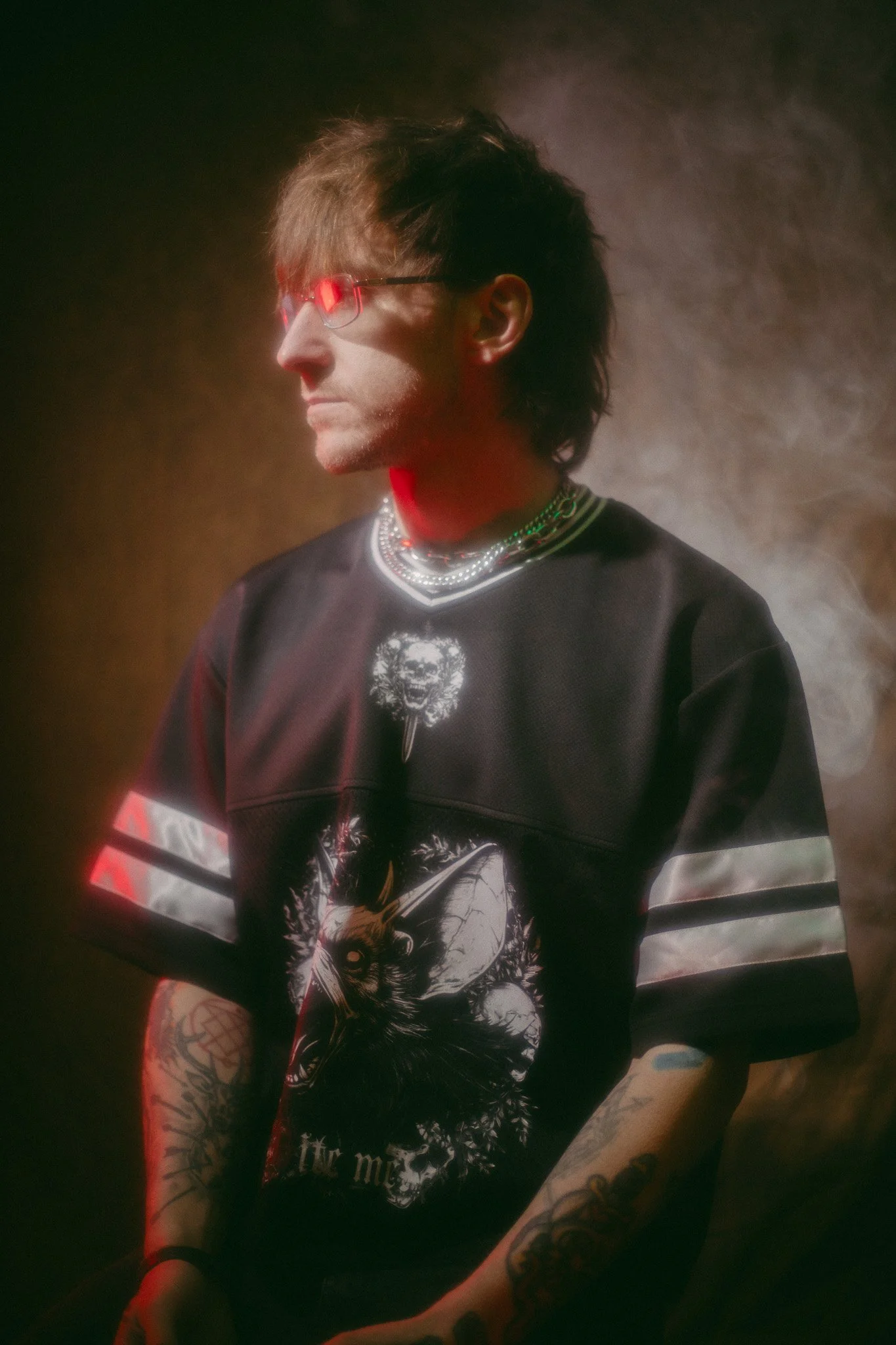 A person with dark, wavy hair wearing glasses with red reflective lenses, a black graphic T-shirt with a wolf graphic, multiple layered necklaces, and visible tattoos on their arm, standing against a dark, smoky background.