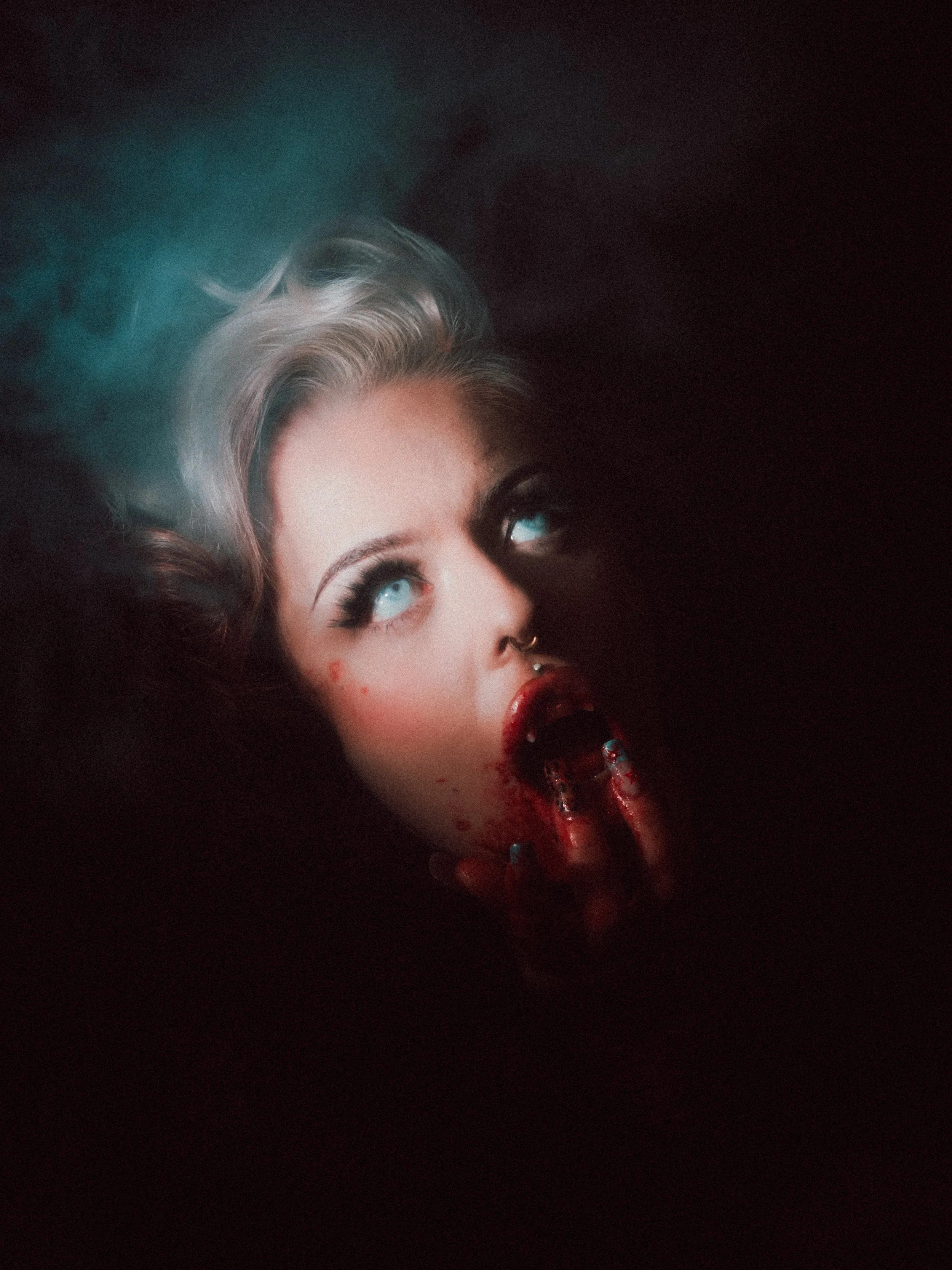 A woman with light-colored hair, bright blue eyes, and makeup with a dark lipstick, lying down in darkness with a blood-stained finger near her mouth and a red lollipop in her mouth.