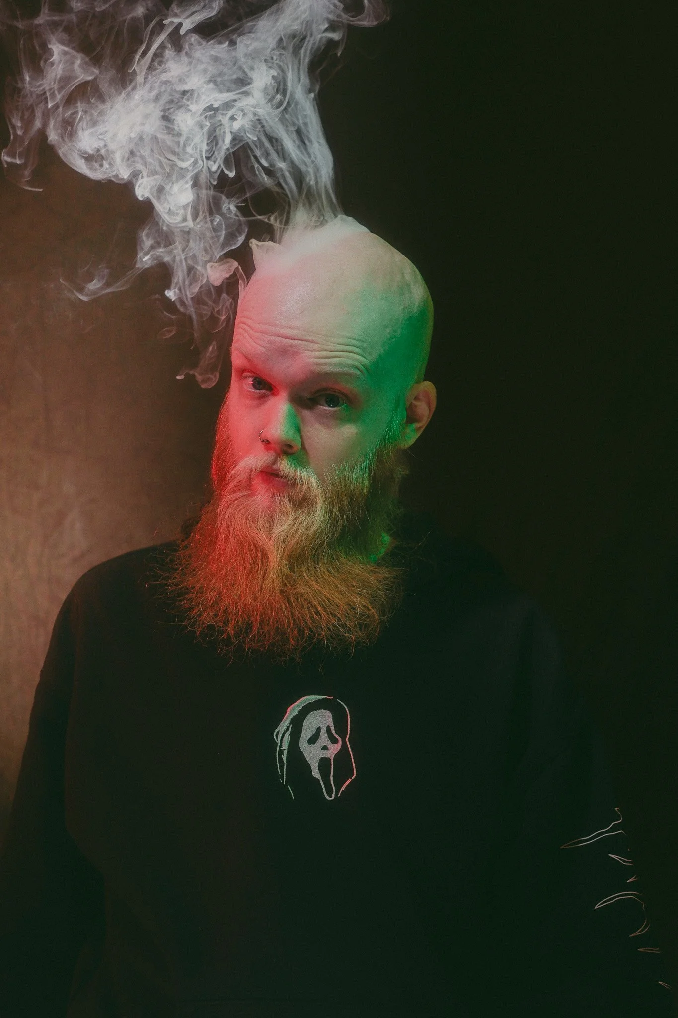 A man with a bald head, red beard, and nose piercing wearing a black shirt with a skull graphic, looking at the camera, with smoke coming from his head against a black background.
