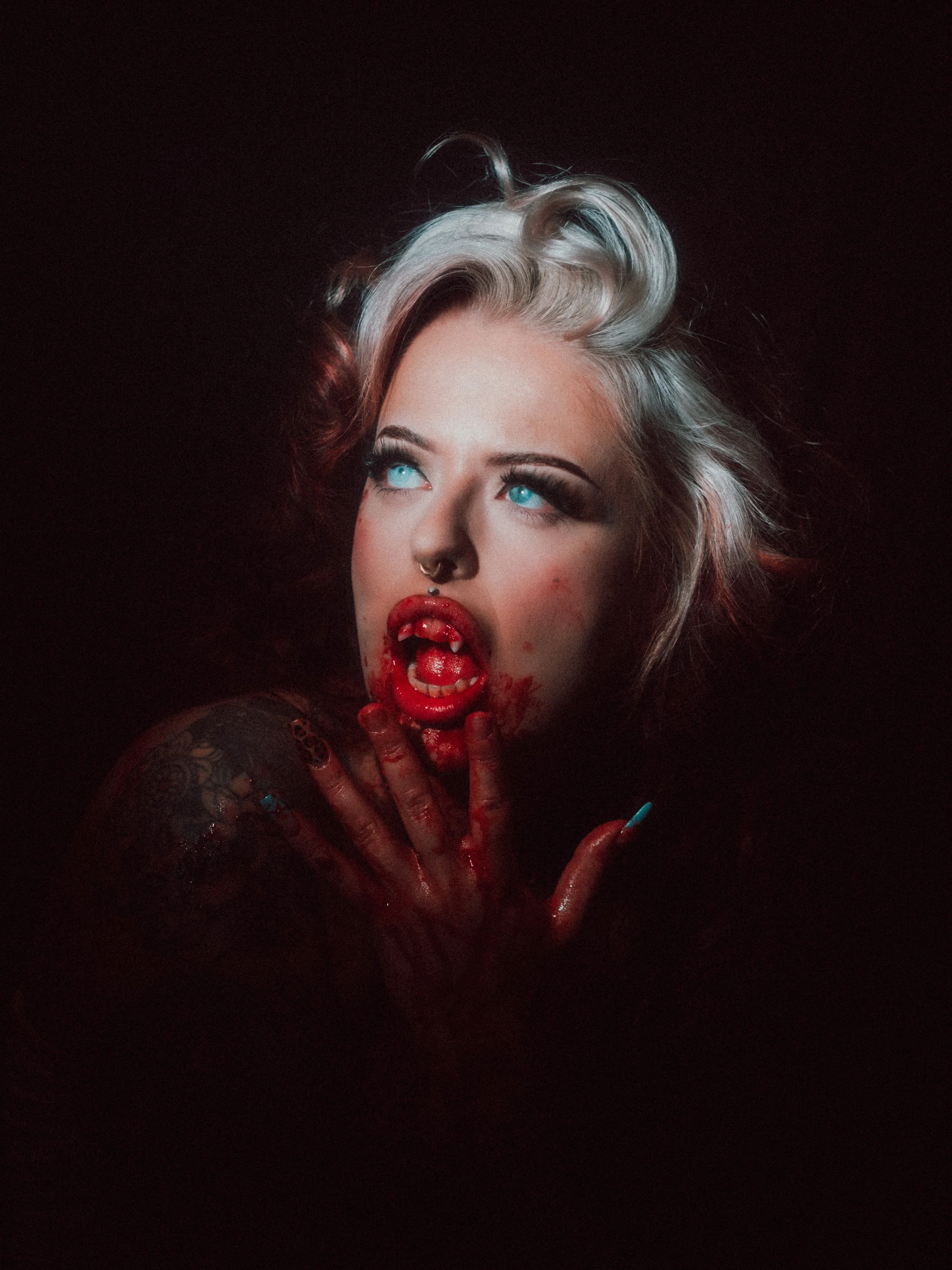 A woman with platinum blonde hair and bright blue eyes has her mouth open, showing sharp vampire fangs. Blood is dripping from her mouth and smeared on her hand, which is near her face, against a dark background.