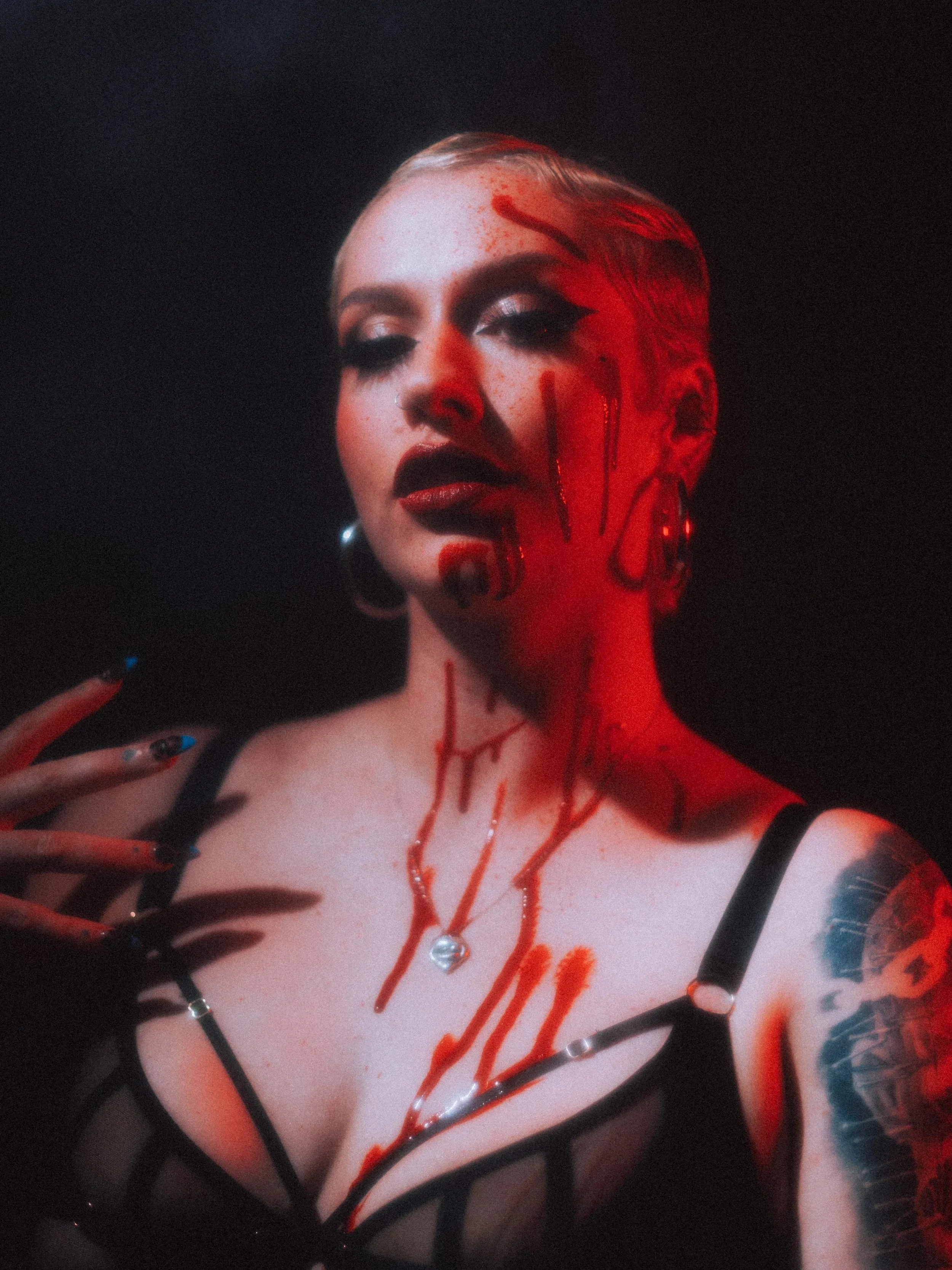 A woman with platinum blonde hair, dark makeup, and red lipstick, dressed in black lingerie, has blood-like red paint on her chest and face. She is wearing a necklace with a pearl pendant and has tattoos on her arm. The background is dark, with red lighting highlighting her face and body.