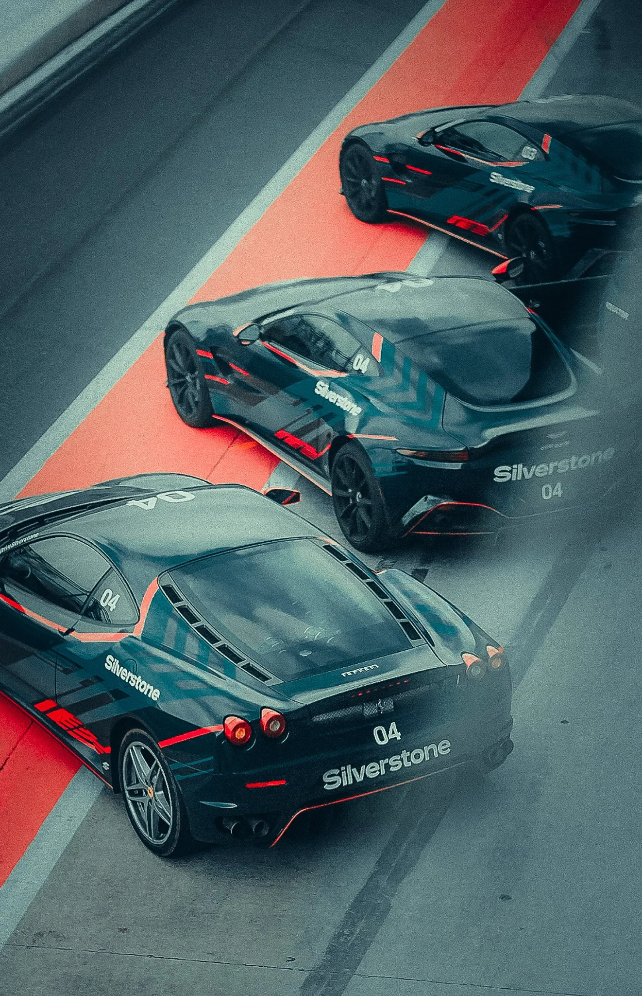 High angle view of two black sports cars with red accents parked on a red parking space next to a gray track, with the word 'Silverstone' and the number '04' on their rear.