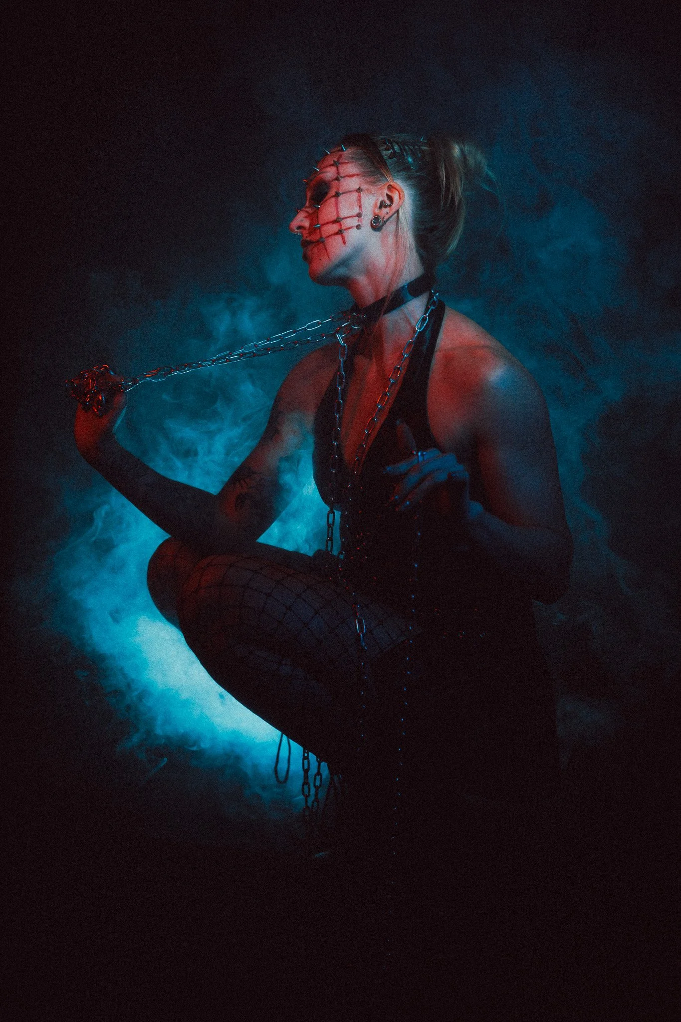 A woman in a dark outfit with chains and spikes, makeup, fishnet stockings, and with face markings, poses in a dimly lit, smoky environment with red and blue lighting.