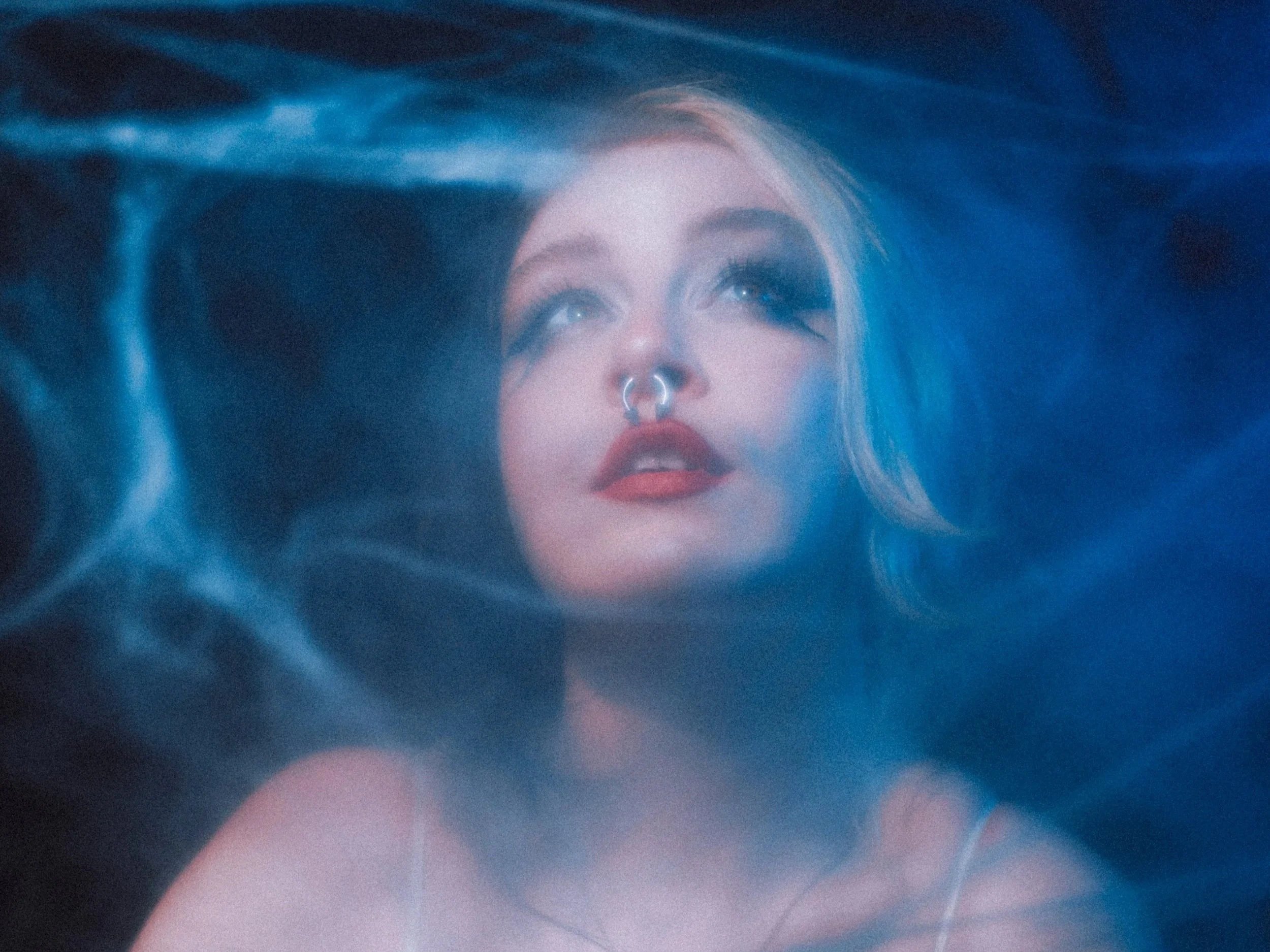 Close-up of a young woman's face with pastel makeup, light hair, and septum piercing, surrounded by a blue and pink dreamy background.