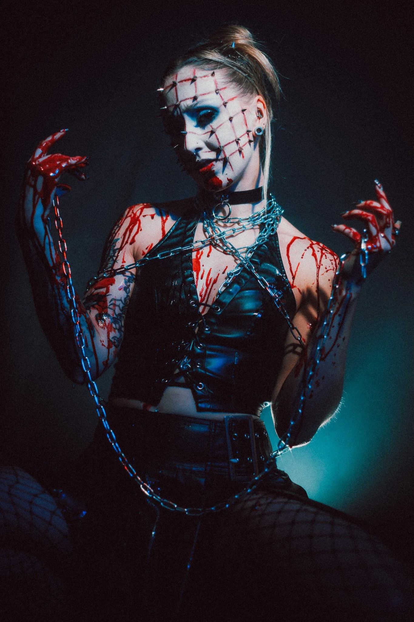 A person with makeup resembling a broken porcelain doll, pierced with safety pins on face, wearing black leather outfit, chains, and fishnet gloves, in front of black background.