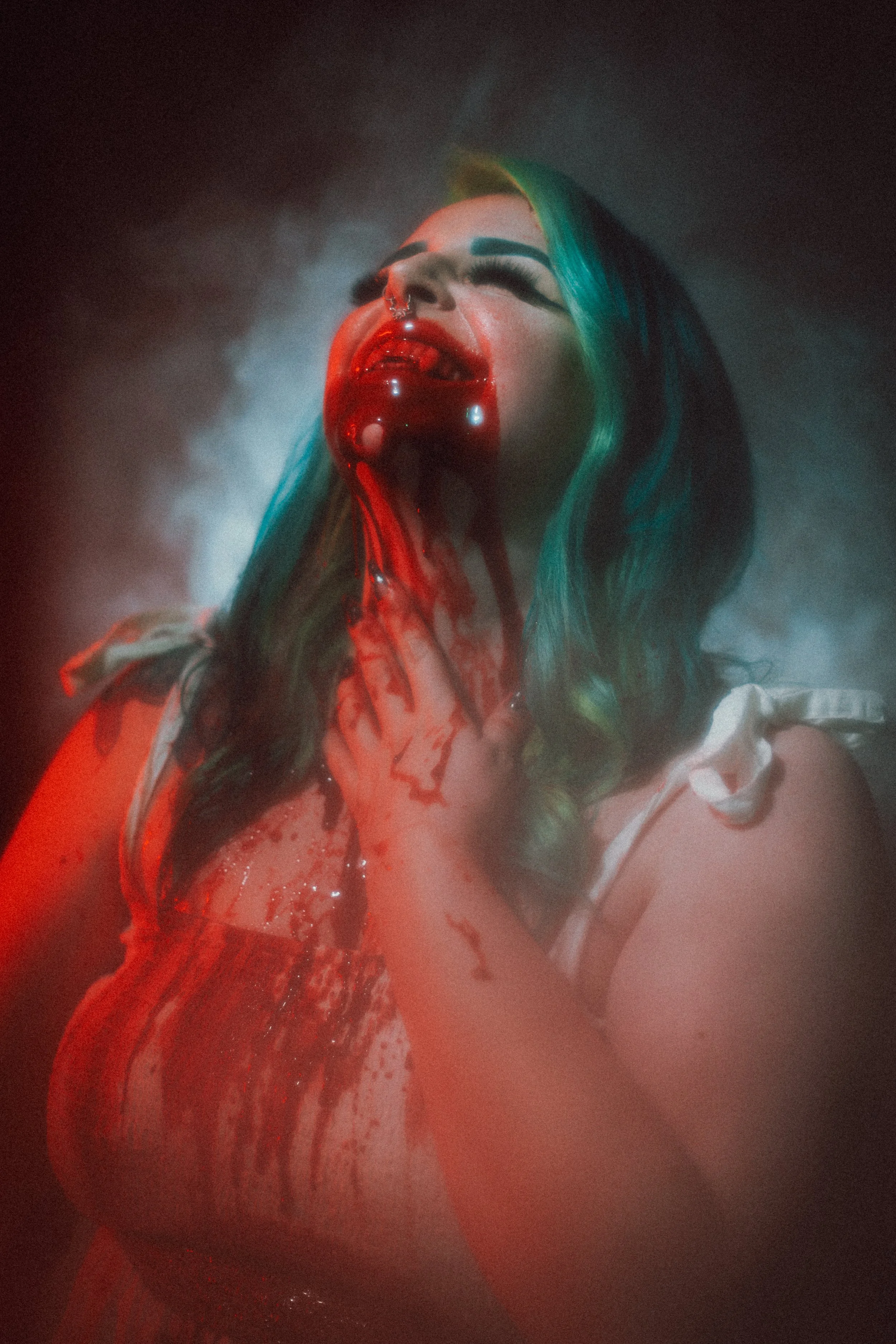 A woman with blue and green hair, makeup, and a nose ring, with her head tilted back and spilling a red liquid from her mouth, creating a blood-like effect.