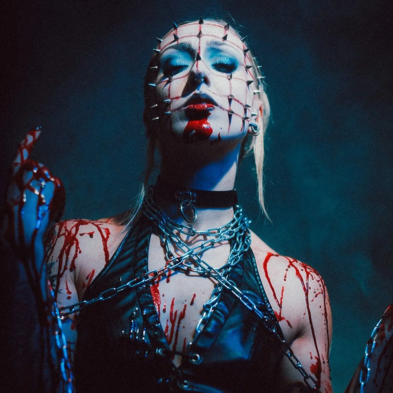 A person with light skin wearing a white mask with spikes and streaks of red, with makeup resembling blood, chains, and a black leather outfit, staring intensely with closed eyes in a dark setting.