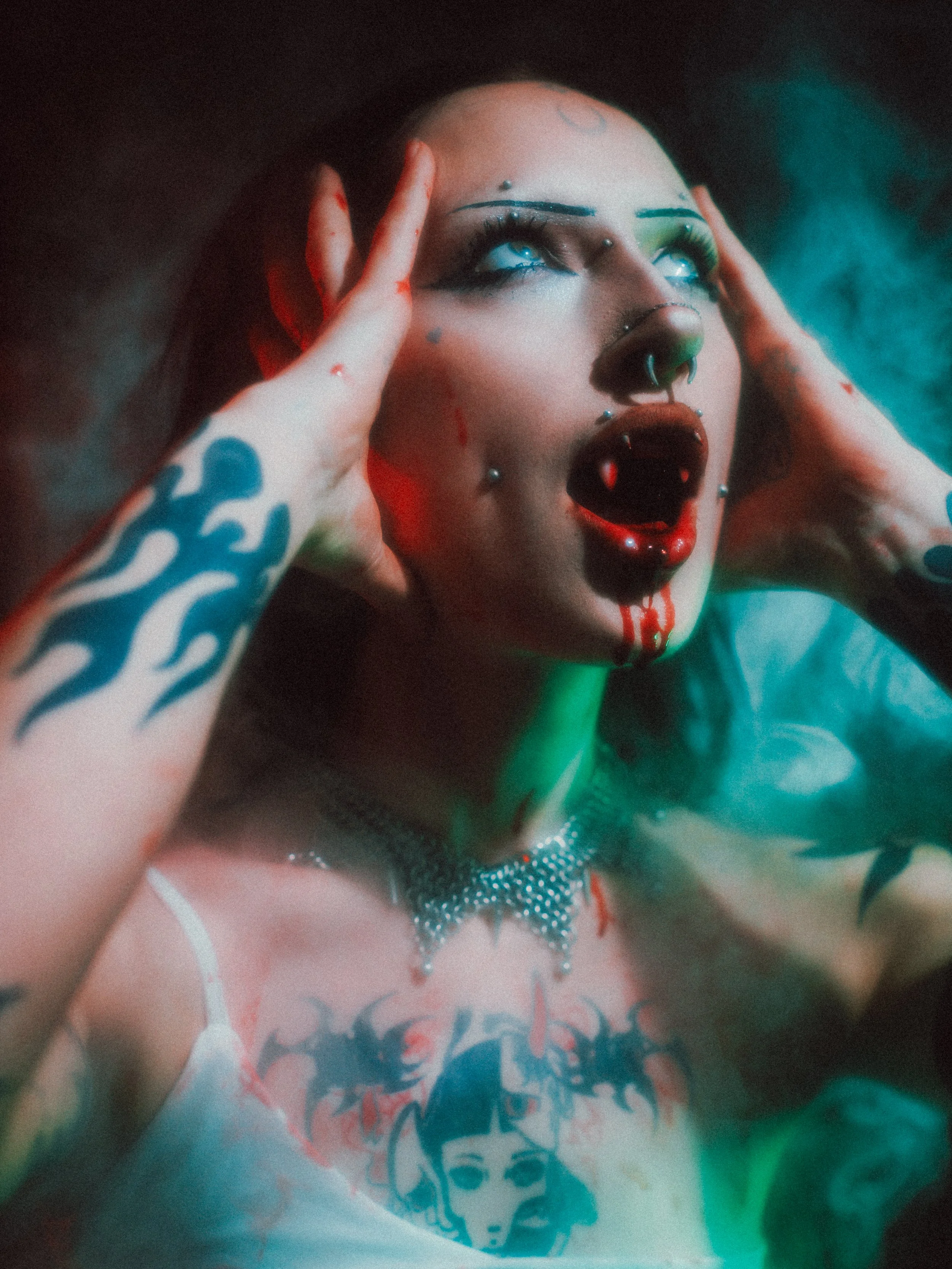 A person with colorful makeup and piercings, holding their head with their hands, wearing a graphic t-shirt with a woman’s face. The photo has dramatic lighting with a mix of red and green hues.