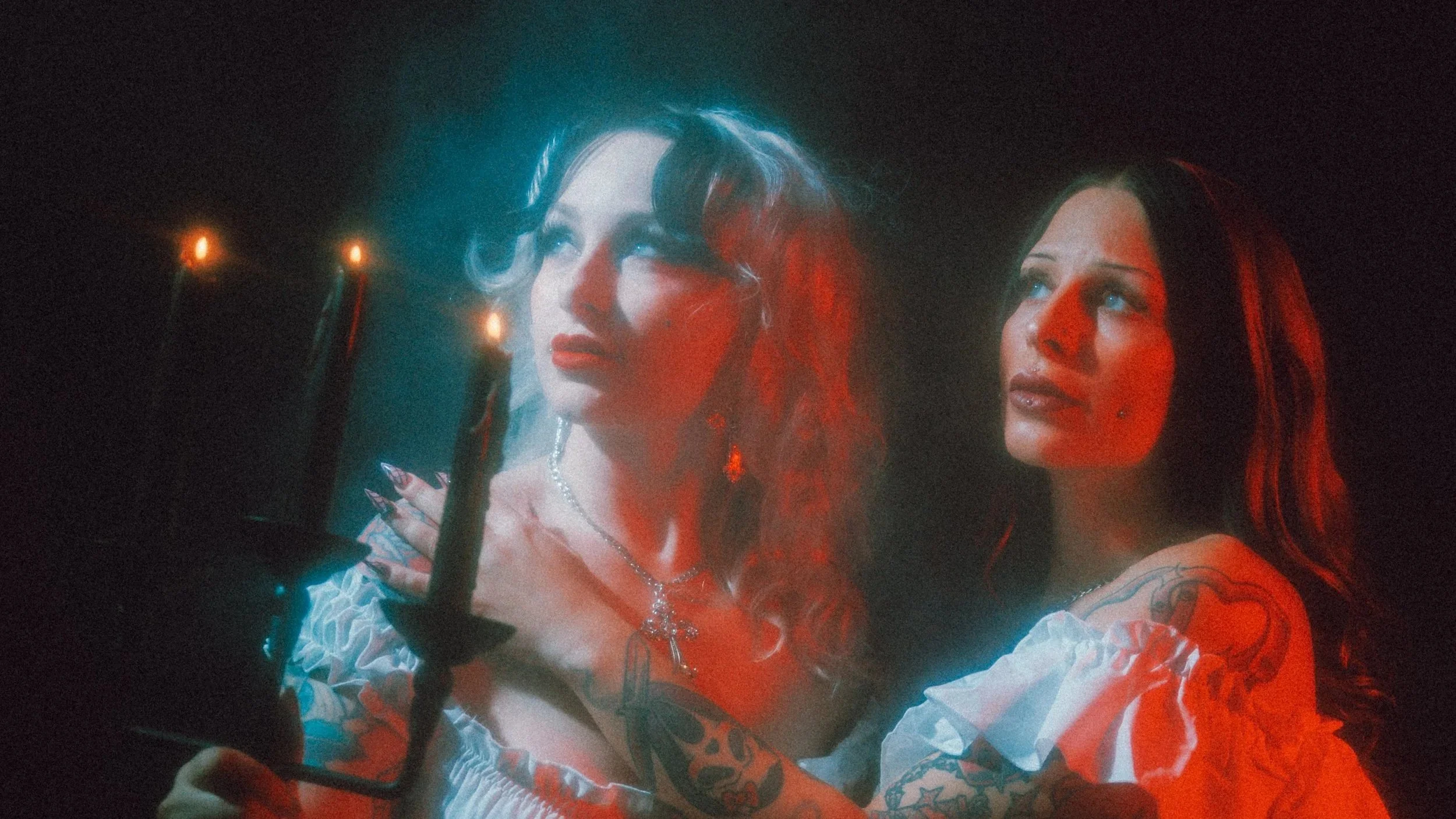 Two women with vibrant hair and tattoos looking into a mirror under red and blue lighting, with candles visible in the background.