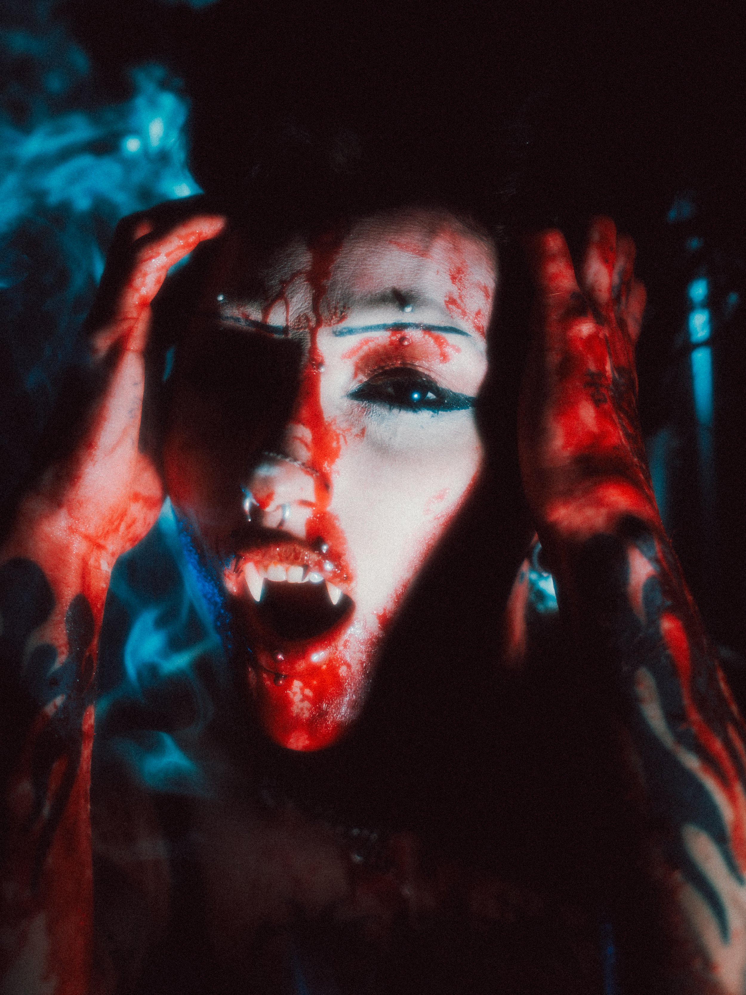 A person with makeup resembling a vampire, with fangs and blood dripping from their mouth and forehead, holding their hair back in a dark setting with eerie blue lighting.