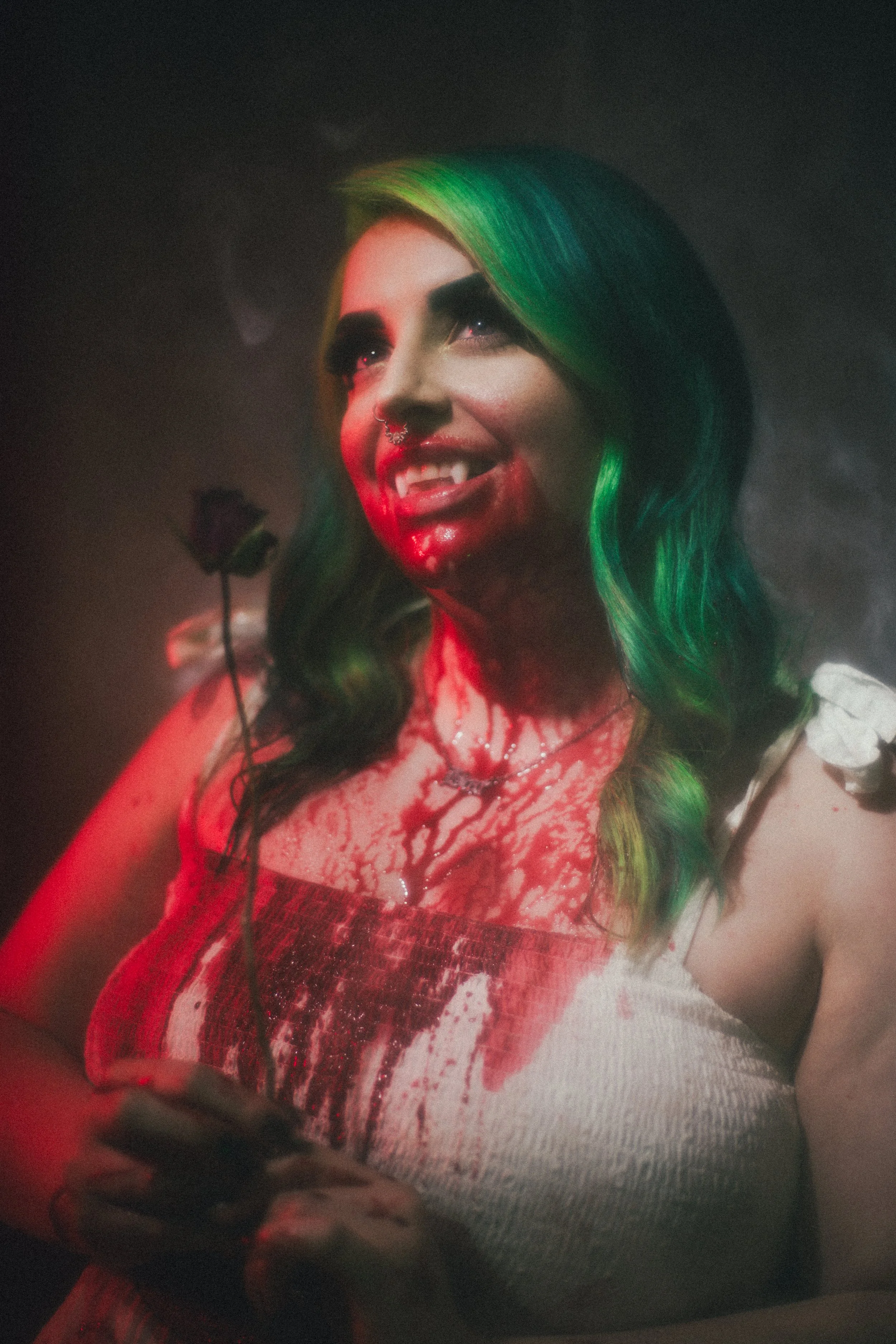 A woman with black and green hair smiling in a dark, foggy setting, wearing a lacy white top with fake bloodstains, a choker, and a septum piercing, holding a small black wand or stick.
