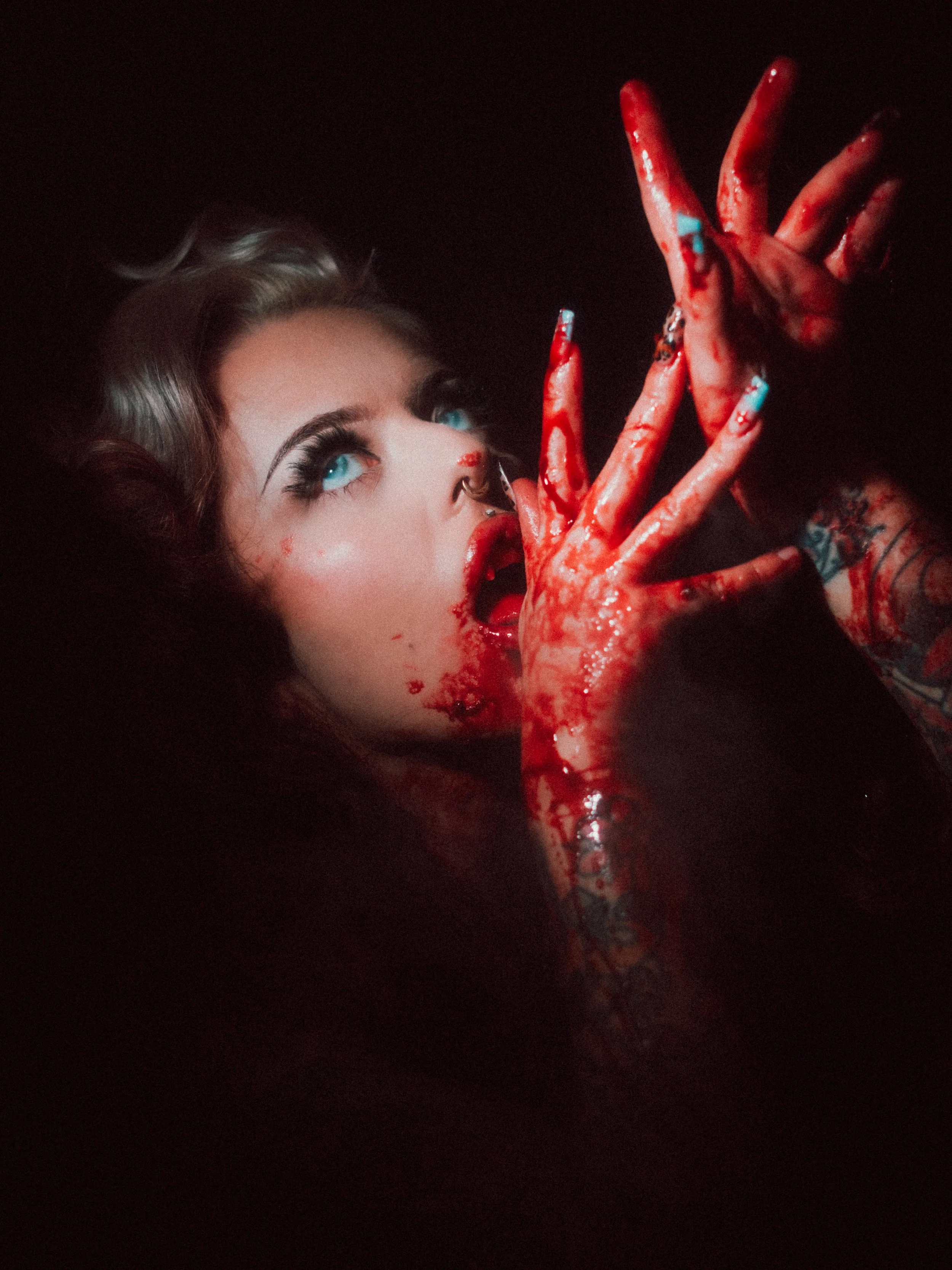 A woman with blue eyes and blonde hair covered in blood, holding blood-covered hands near her face against a dark background.