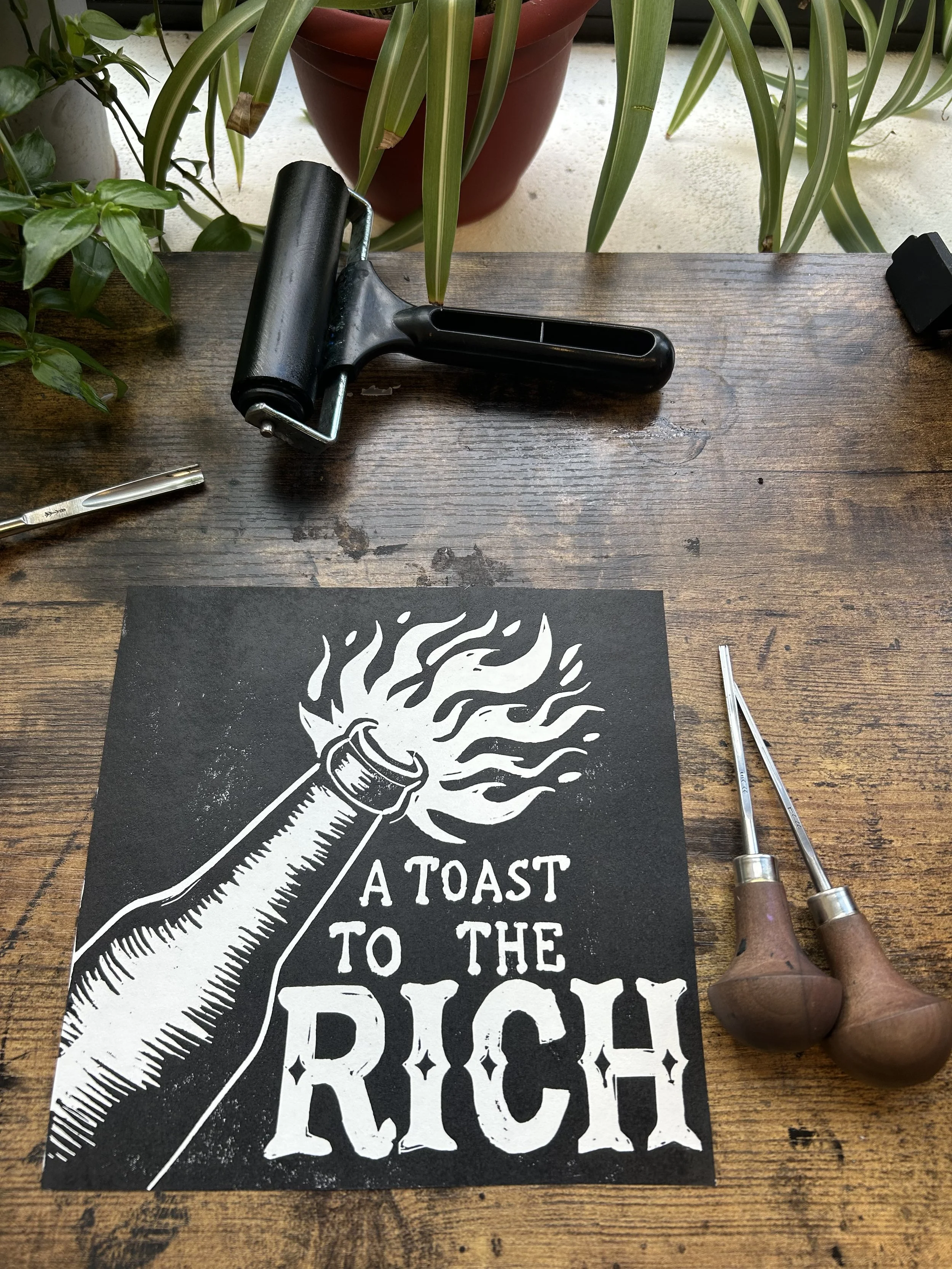A linoleum block printing project with a black and white design of a spray can emitting flames, accompanied by the phrase "TOAST TO THE HIGH REACH" in bold text, on a wooden table with printmaking tools and surrounding plants.