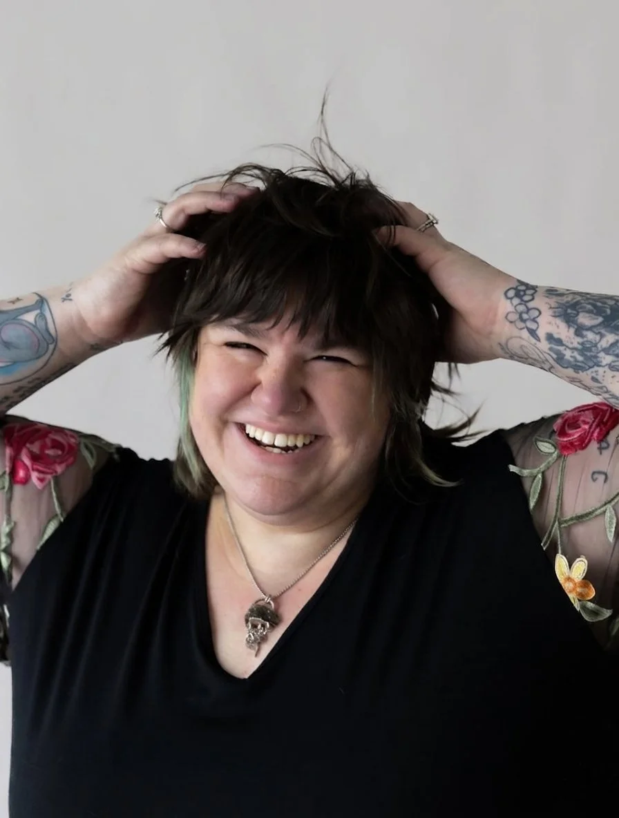 A woman with short, dark hair and tattoos on her arms, smiling happily with her hands in her hair, wearing a black top with floral embroidered sleeves and a necklace with a skull pendant.