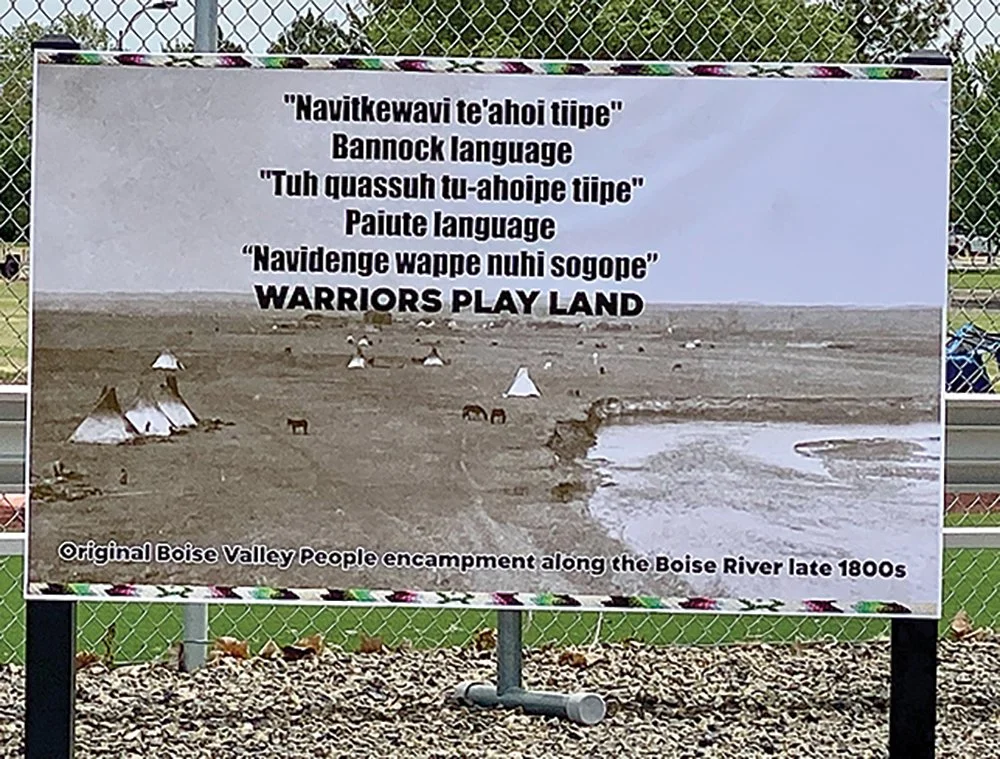 Historical photograph of Boise Valley Native People encampment along the Boise River from the late 1800s, with a quote in Bannock and Paiute languages about playing land, on a display board outside.