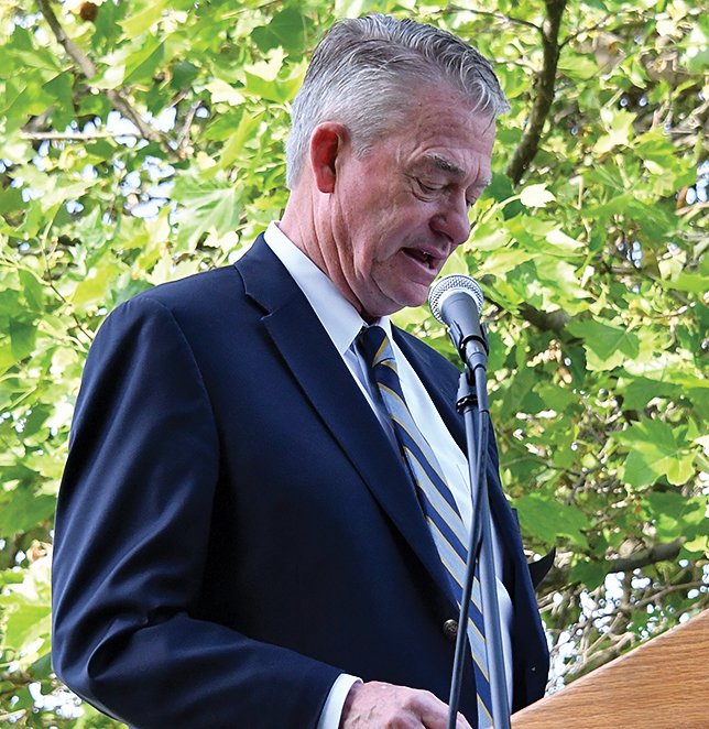 An older man in a dark suit, white shirt, and striped tie speaking at a podium with a microphone, outdoors with green foliage in the background.