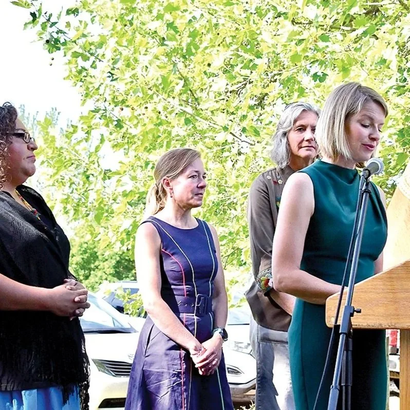 Four women standing outdoors under a tree, with one woman speaking at a wooden podium and microphone, while the others listen.