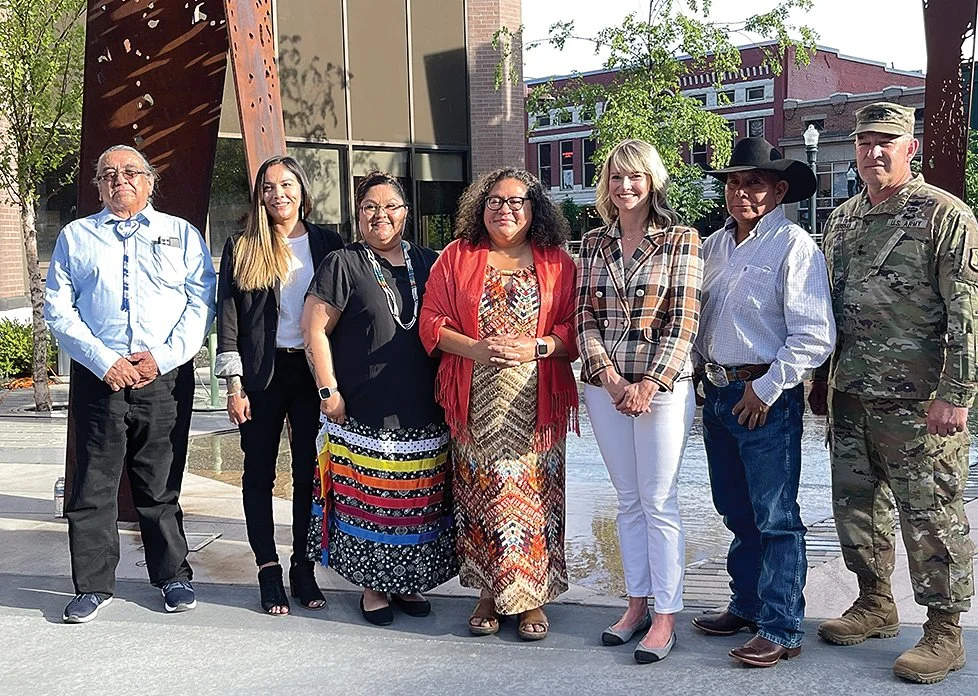 Group of seven diverse people standing outdoors near a reflective building and trees.