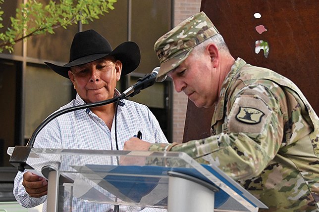A military officer in camouflage uniform signing an outdoor document or plaque while a man in a cowboy hat and striped shirt looks on.