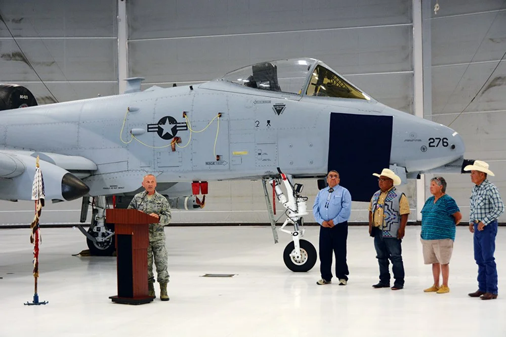 A military person standing at a podium in front of a large fighter jet inside an aircraft hangar, with four civilians standing to the right of the jet.