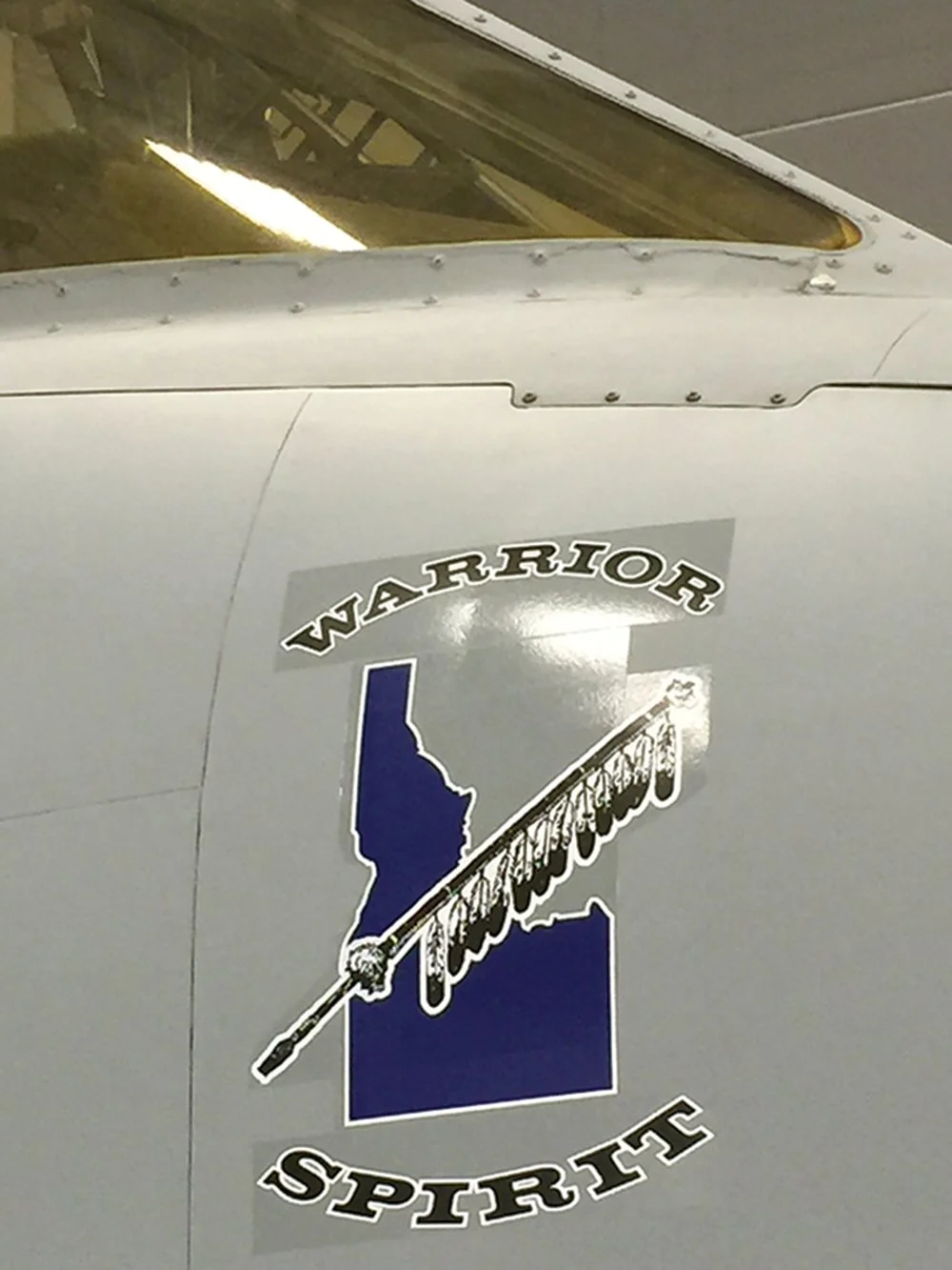 Close-up of an aircraft's fuselage with a sticker featuring a blue silhouette of the state of Idaho, a sword, and the words "WARRIOR SPIRIT" and "GUNSMITHING".