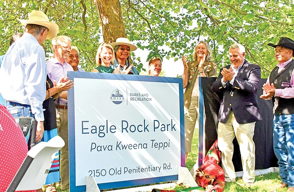 Group of people gathered outdoors for a celebration at Eagle Rock Park, with a large sign that reads 'Eagle Rock Park, Pava Kweena Teppi, 2150 Old Penitentiary Rd.' They are smiling, clapping, and dressed in casual and semi-formal attire, with some wearing hats.