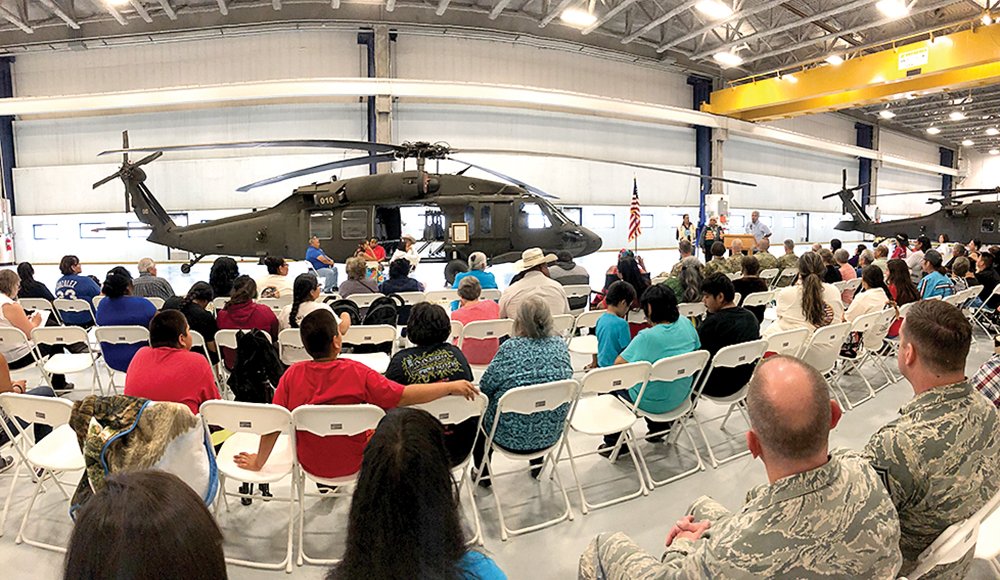 People seated in chairs inside a hangar, viewing two military helicopters and a presentation or demonstration.