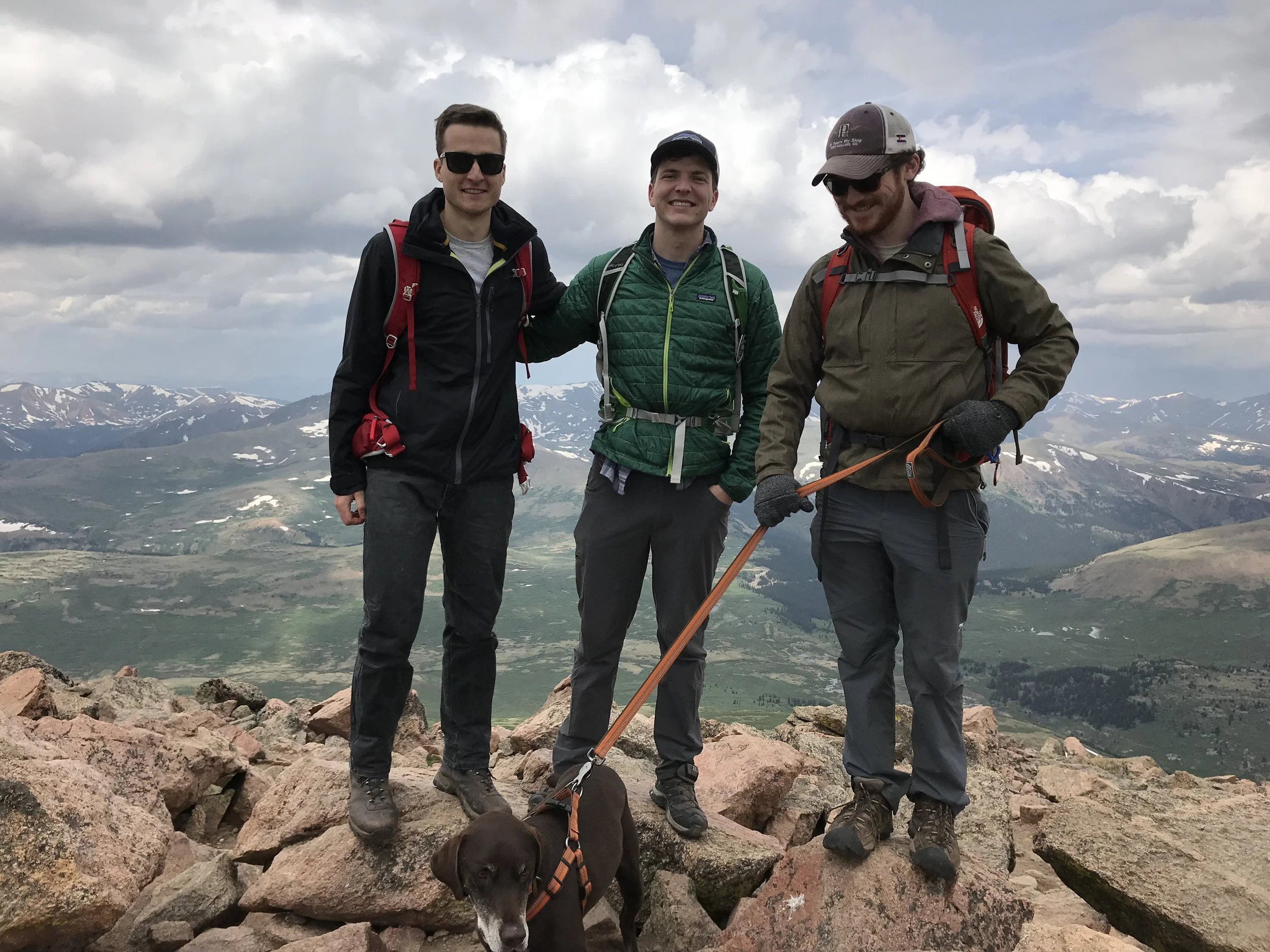 Three young men dressed in hiking gear standing on a mountain with rocky terrain, with a scenic view of snow-capped mountains and cloudy sky in the background. A black dog with a harness is lying on the rocks in the foreground.