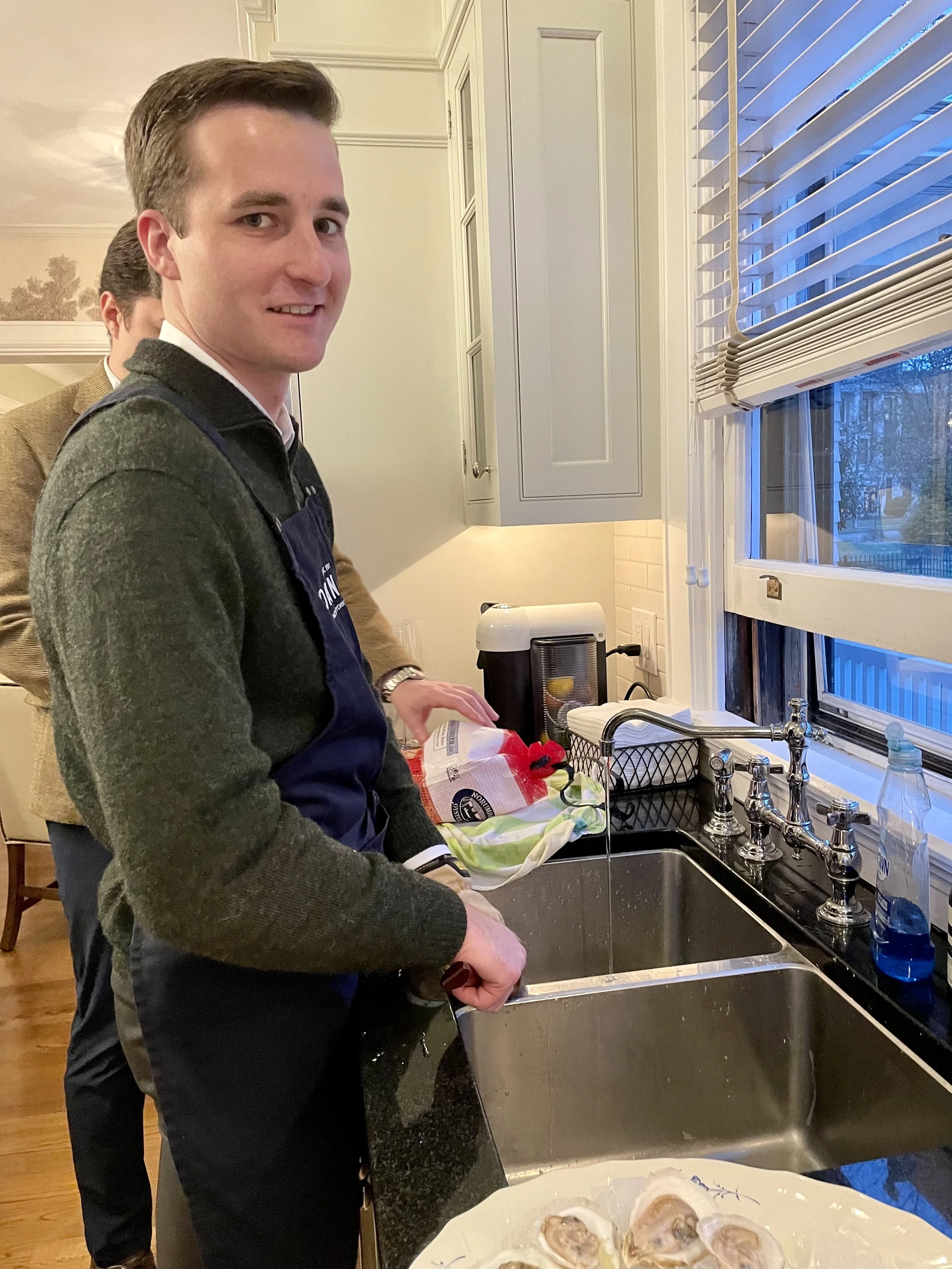 A young man with brown hair and a smile, wearing a dark apron, washing oysters in a kitchen sink. There is a window with horizontal blinds and another person in the background.