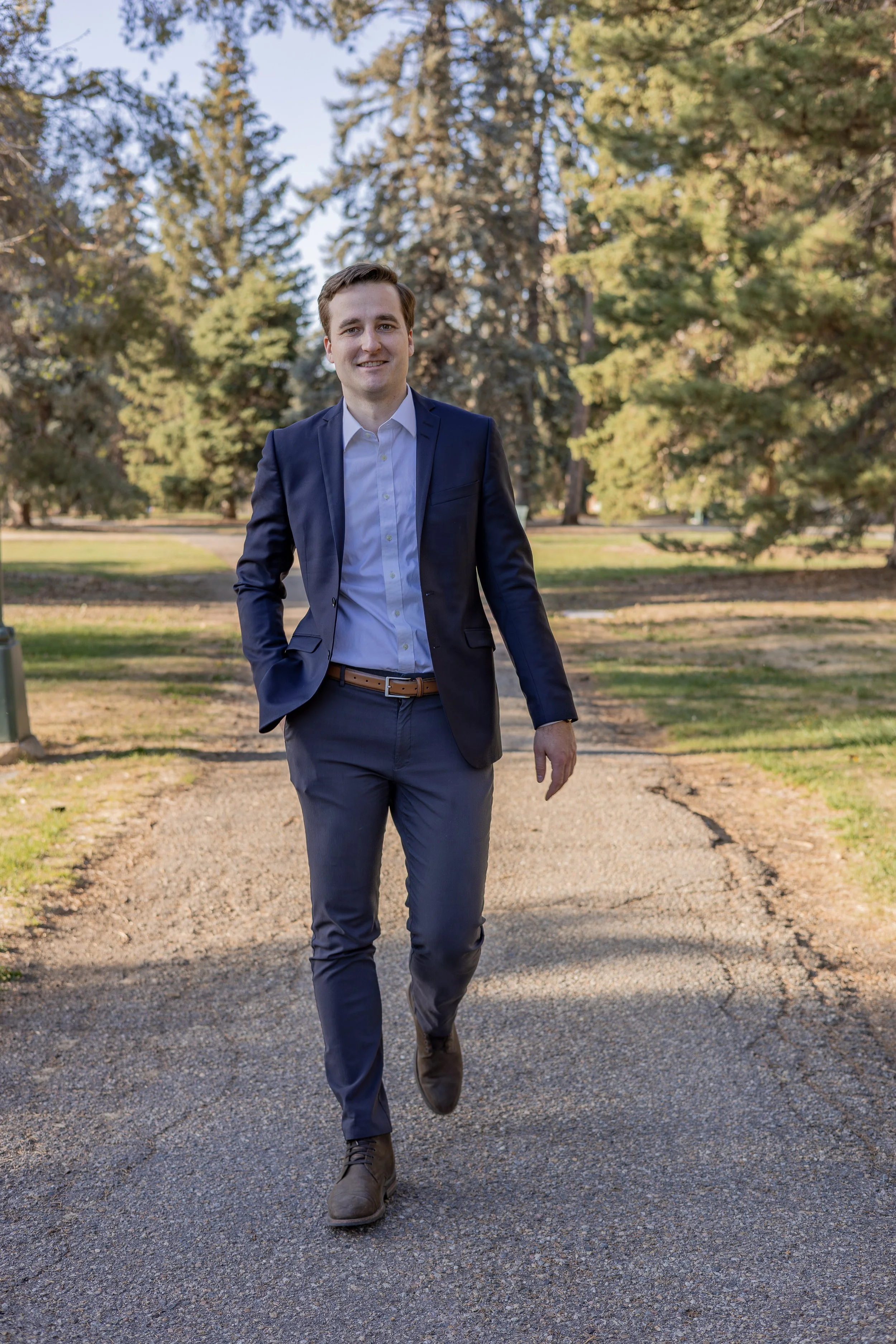 A man in a navy blue suit and white shirt walking on a gravel path in a park with green and yellow trees.