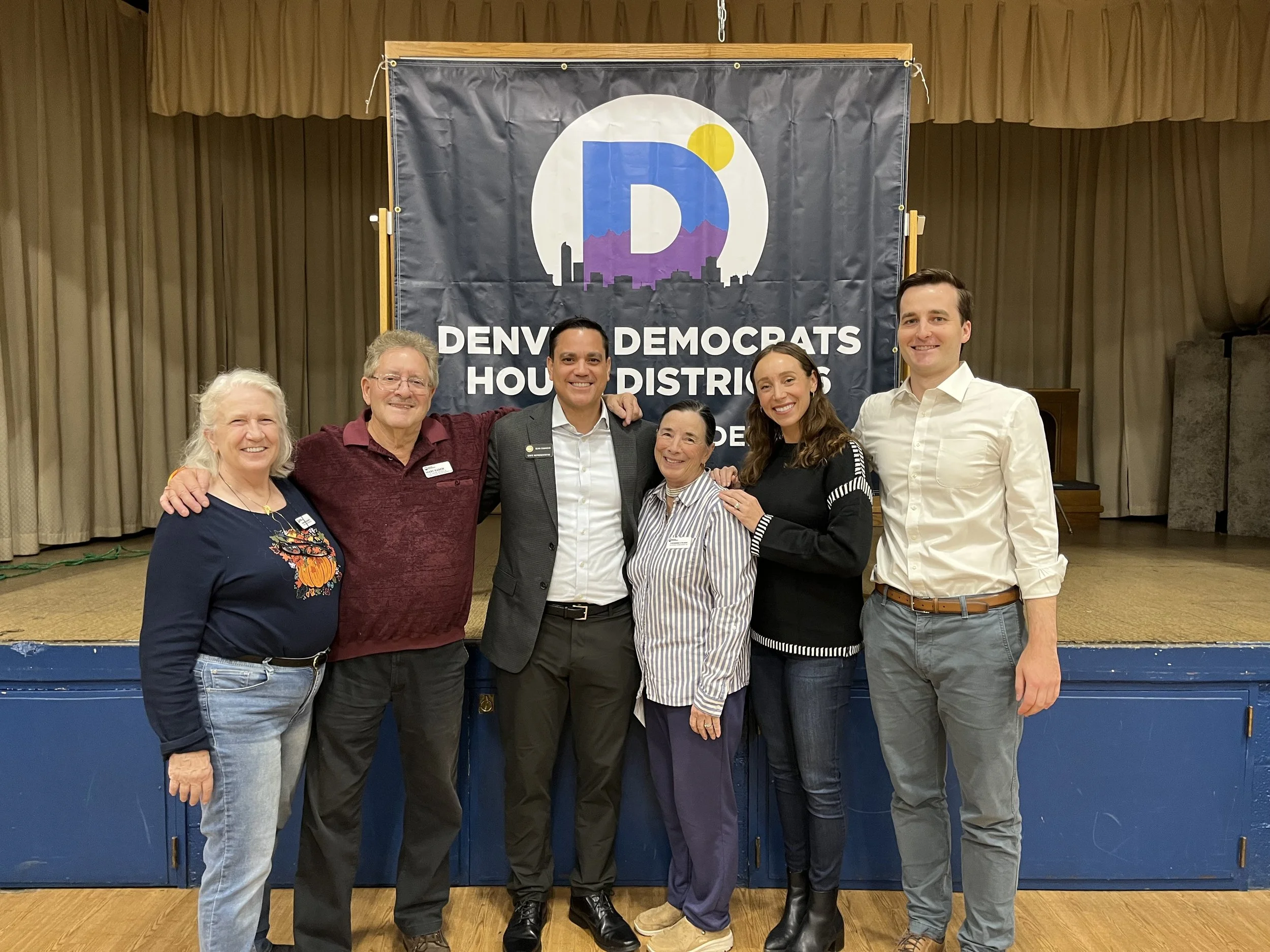 A group of six diverse people standing together in a room with a stage and a large banner that reads 'Denver Democrats House Districts 6' They are smiling and posing for a photo.