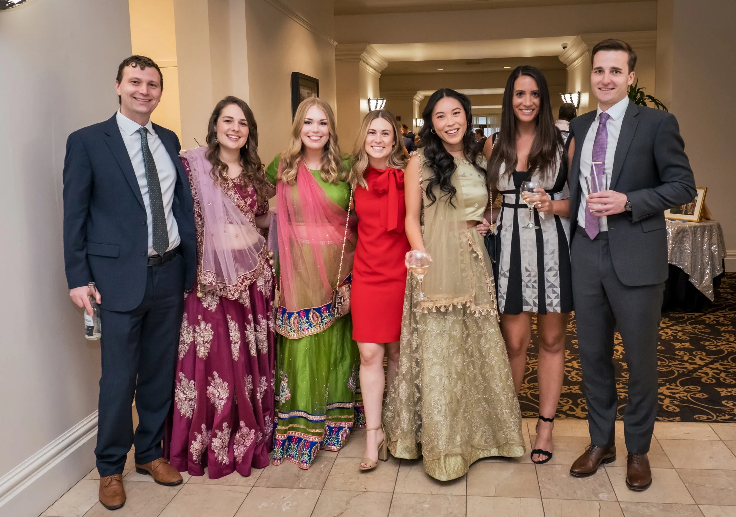 A diverse group of seven people dressed in formal and traditional attire at an indoor event, standing in a line and smiling at the camera.
