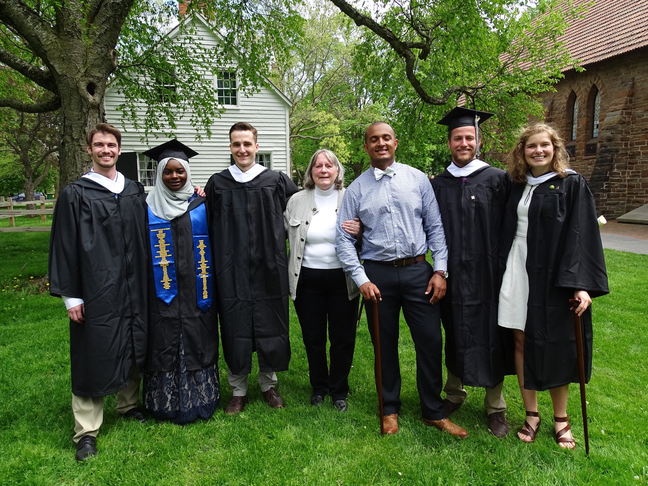 Group of six people, including graduates in caps and gowns, posing outdoors under a large tree with green leaves and a historic building with brick and white siding in the background.