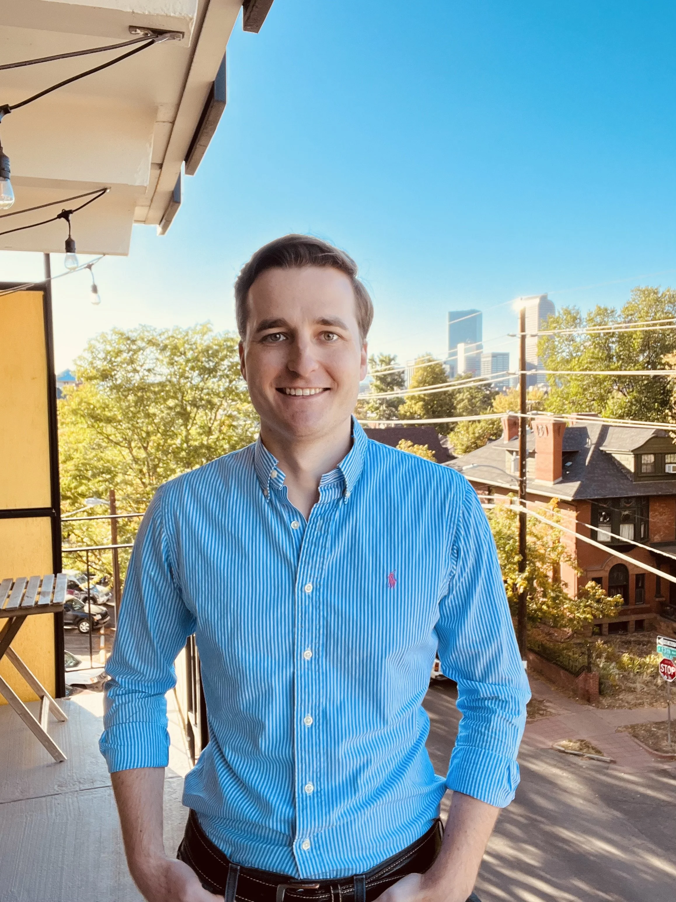 A man with light skin and short brown hair wearing a light blue striped button-up shirt, standing outdoors on a balcony with a cityscape, trees, and a bright blue sky in the background.