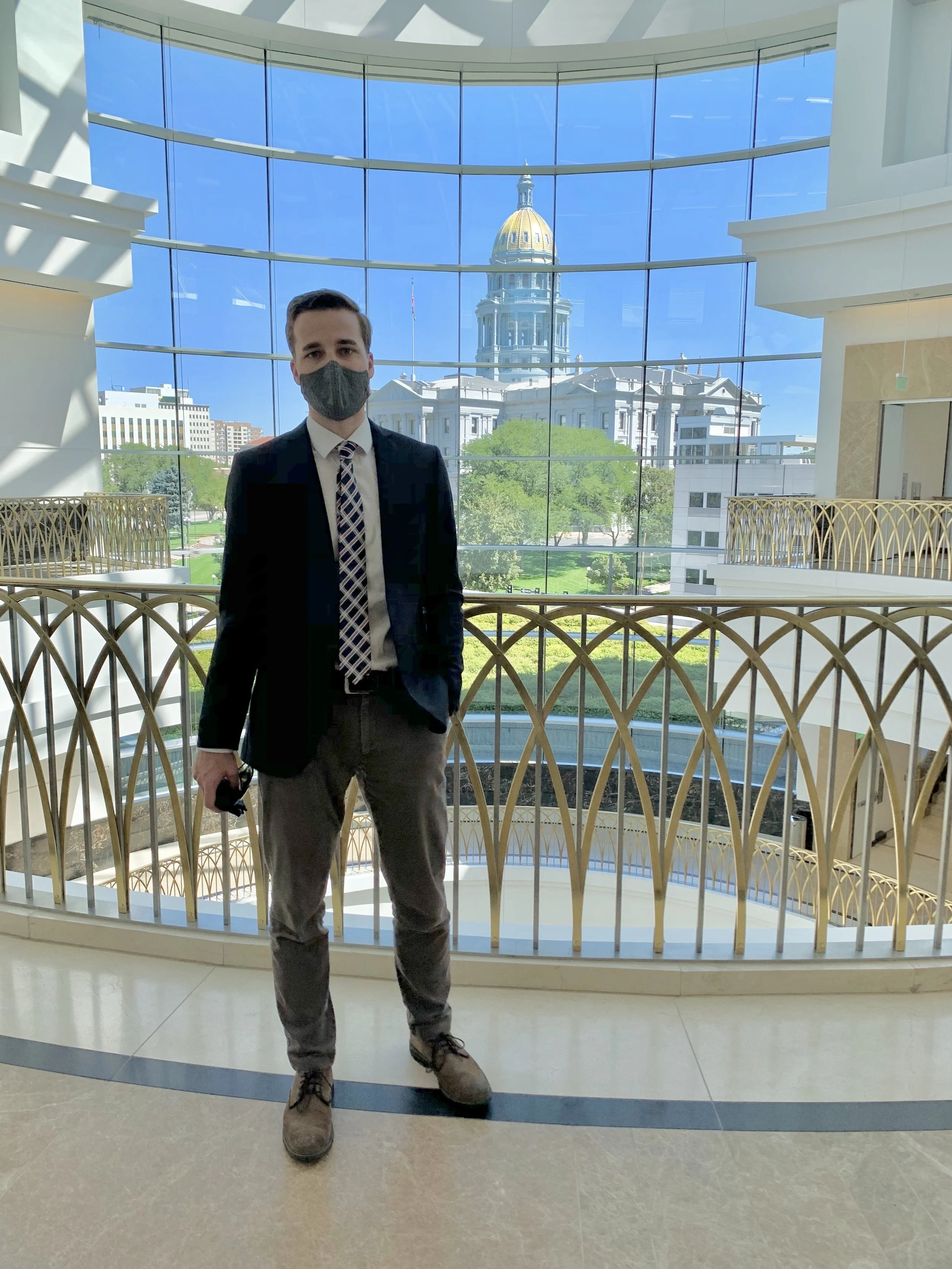 A man dressed in a business suit with a patterned tie and wearing a face mask stands inside a modern building with large glass windows, with a view of a historic building with a domed roof outside.