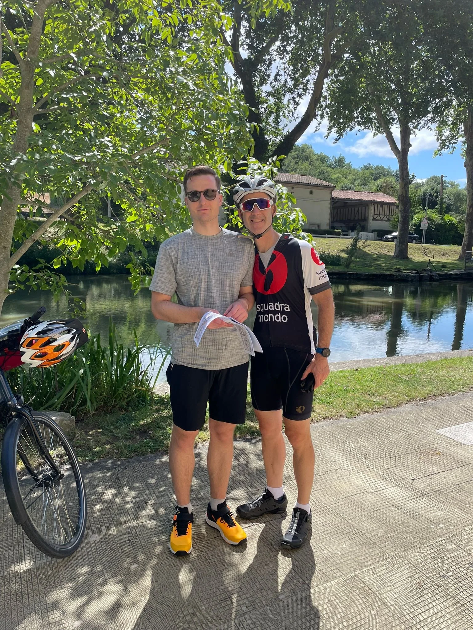 Two men are standing near a body of water with green trees and houses in the background. The man on the left is in casual clothing, and the man on the right is wearing cycling gear, including a helmet and sunglasses. A bicycle helmet is resting on a 