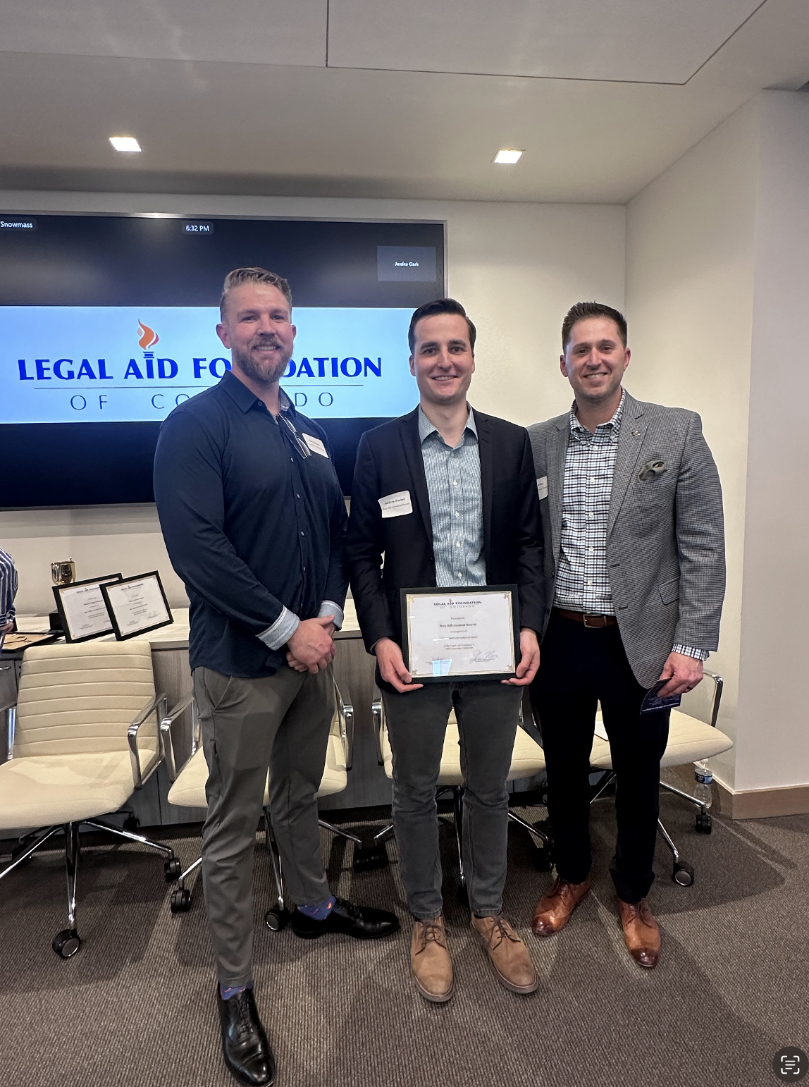 Three men in business attire standing together during an award ceremony, with one holding a certificate. Behind them is a large screen with the logo of the Legal Aid Foundation of Colorado.